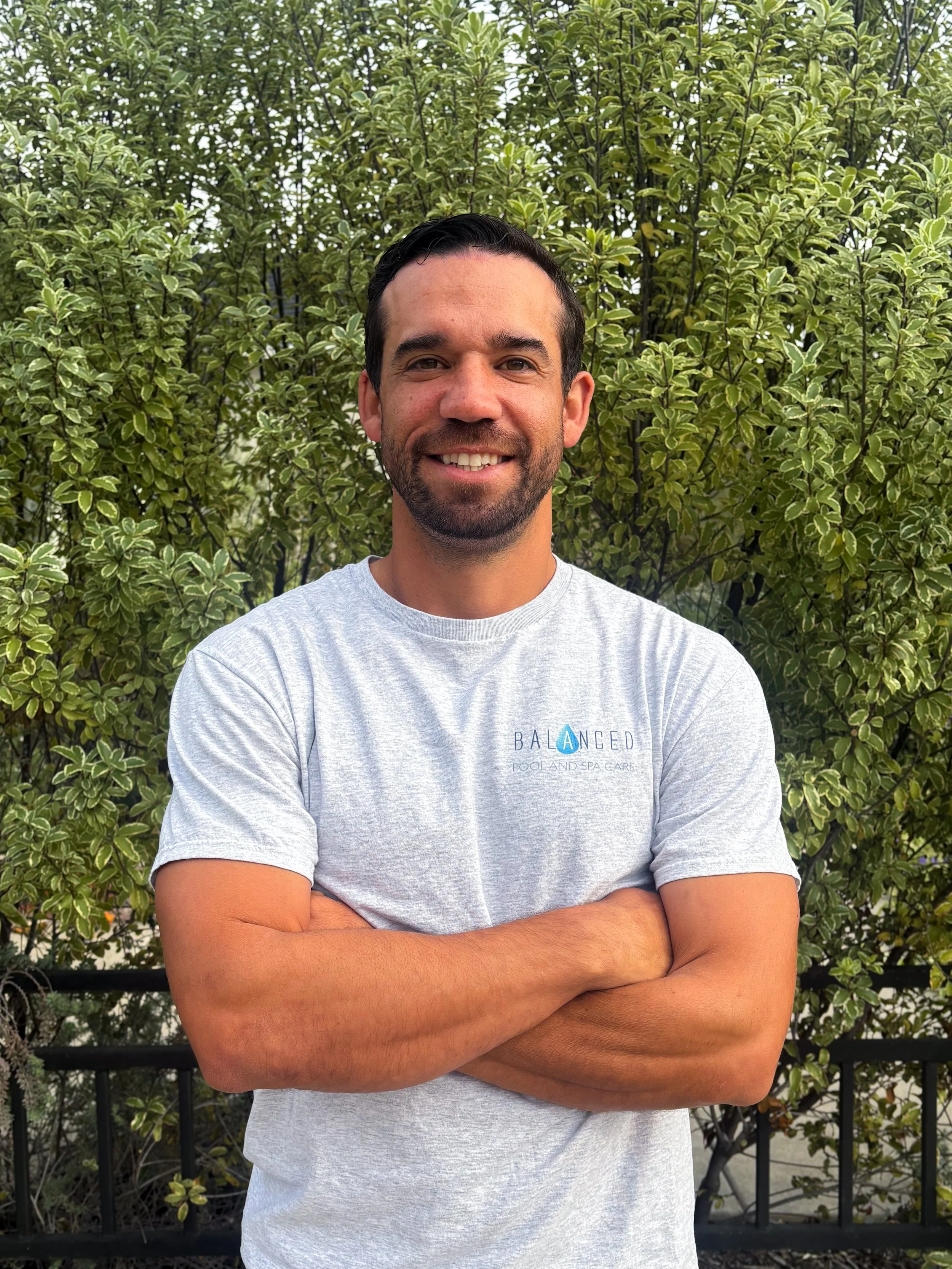 image of owner, devin garcia, standing with hands folded crossed in front of greenery, wearing a grey Balanced Pool and Spa Care Shirt
