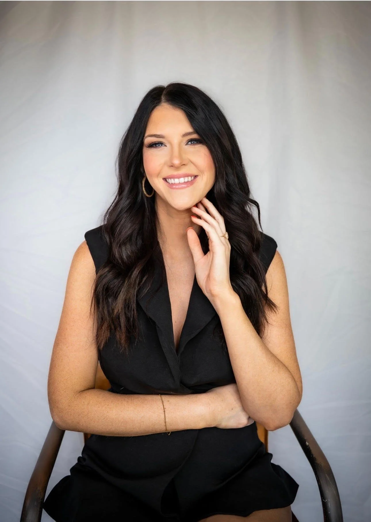 A smiling woman with long dark wavy hair, wearing a sleeveless black top and gold hoop earrings, sitting with her hand resting on her chin against a plain light-colored background.