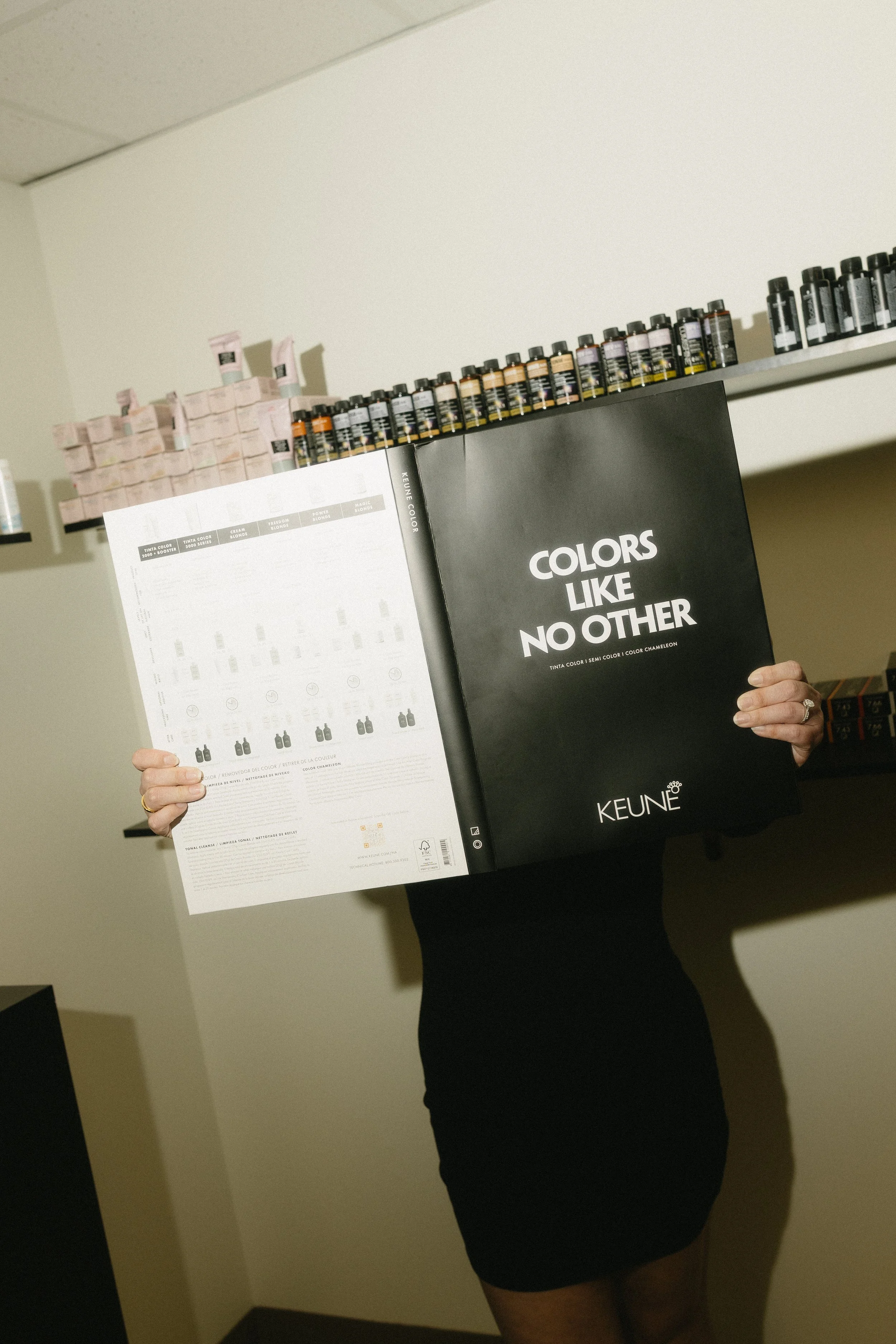 Person holding a catalog titled "Colors Like No Other" in front of shelves stocked with various bottles of ink or paint.