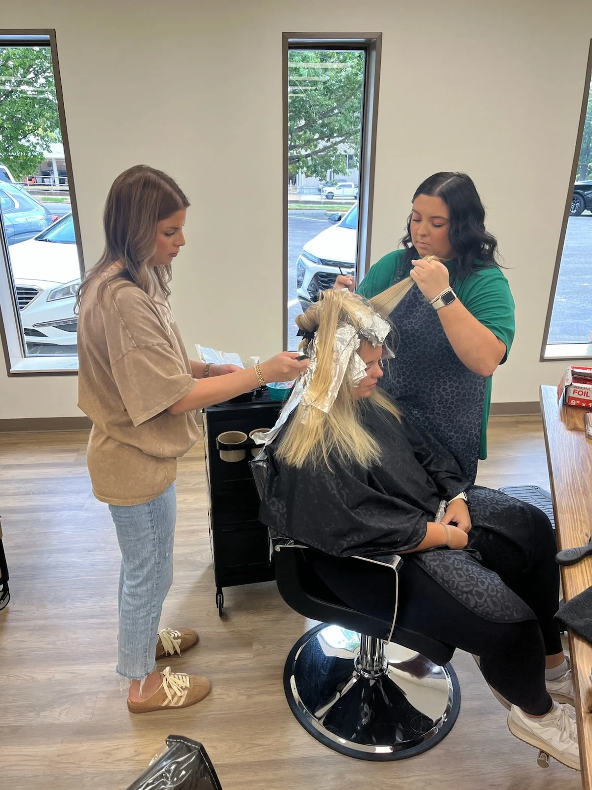 A woman with blonde hair sitting in a salon chair getting her hair colored, while a stylist applies hair dye and another assistant assists nearby.