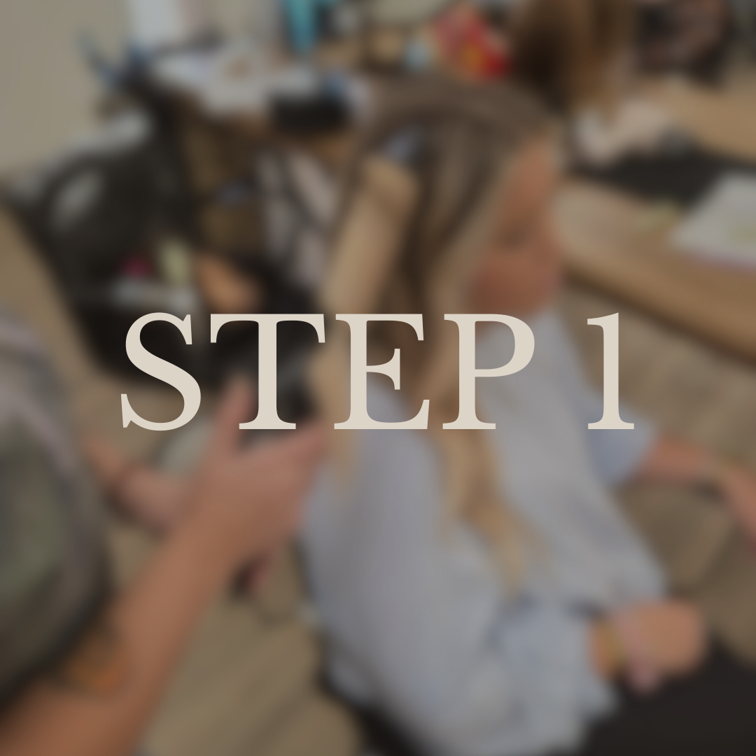 A young girl with blonde hair sitting at a desk while someone brushes her hair