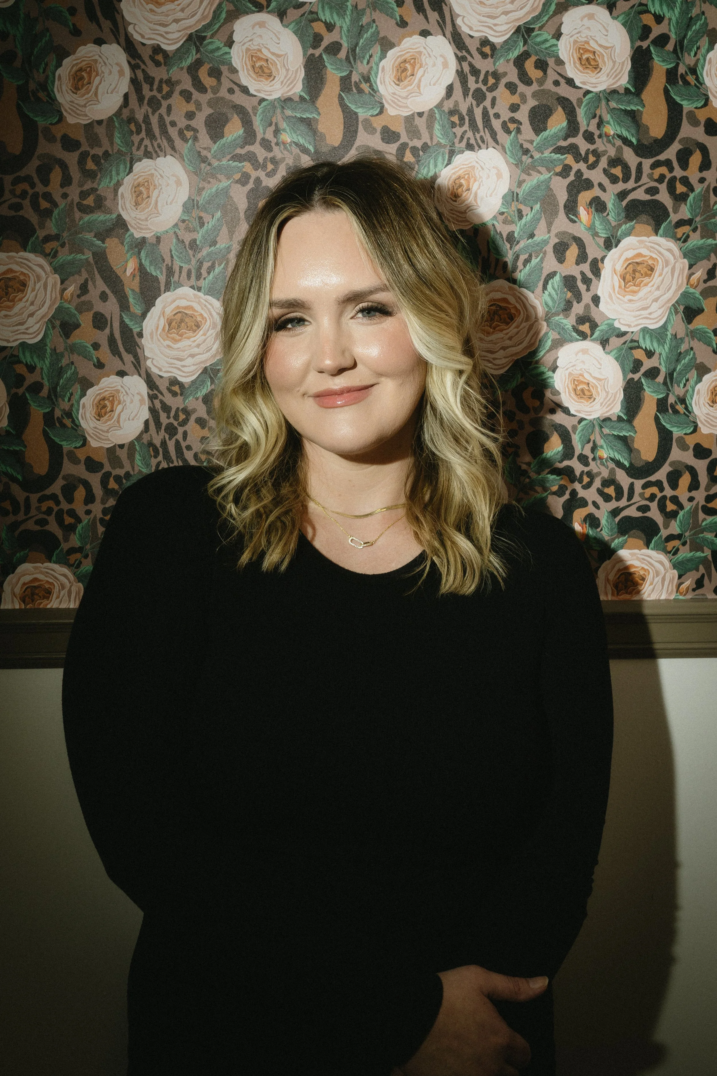 A woman with blonde, wavy hair smiles while posing in front of a floral and animal print wallpaper background.