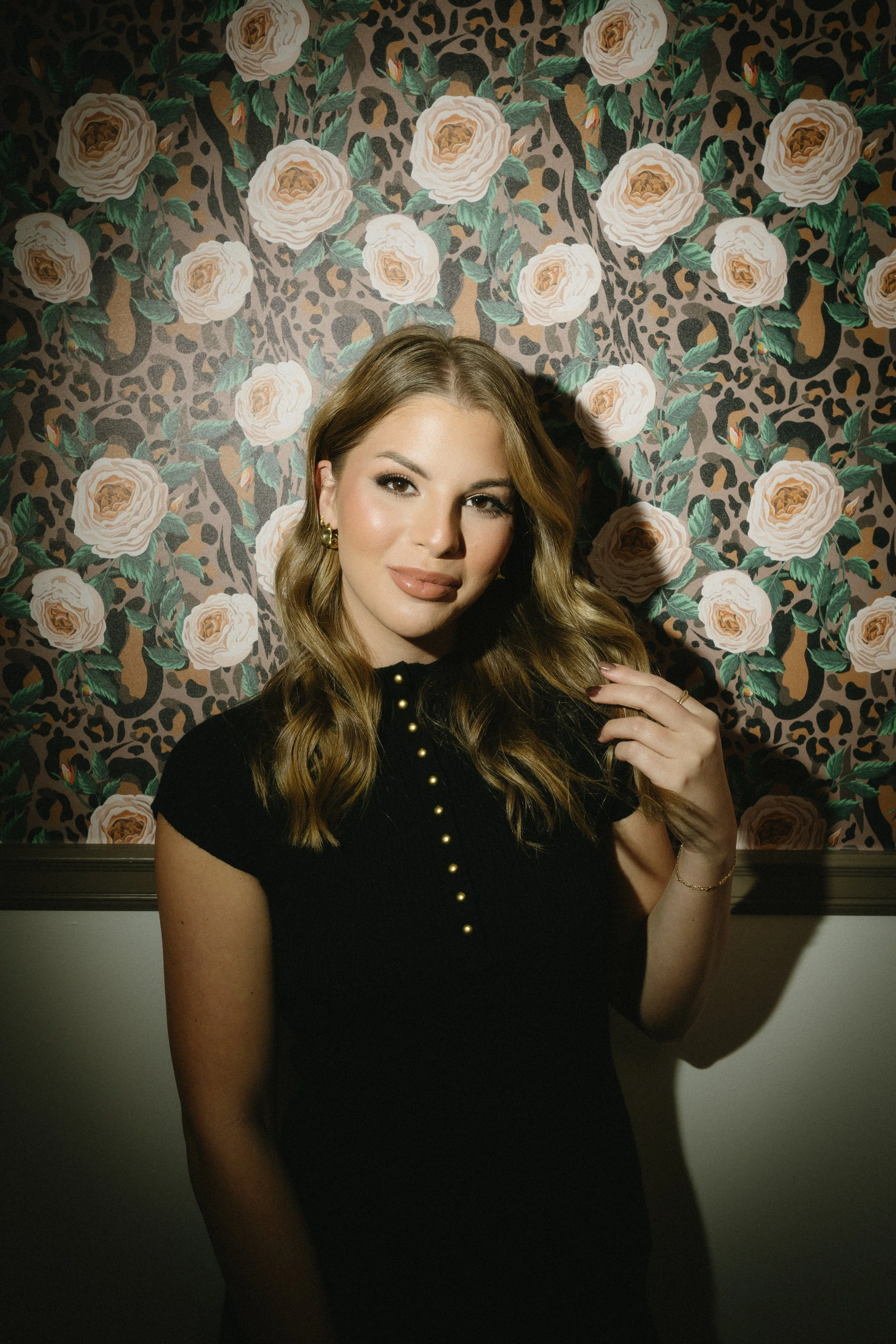 A woman with wavy light brown hair and makeup, wearing a black top with gold buttons, standing in front of floral and animal print wallpaper.