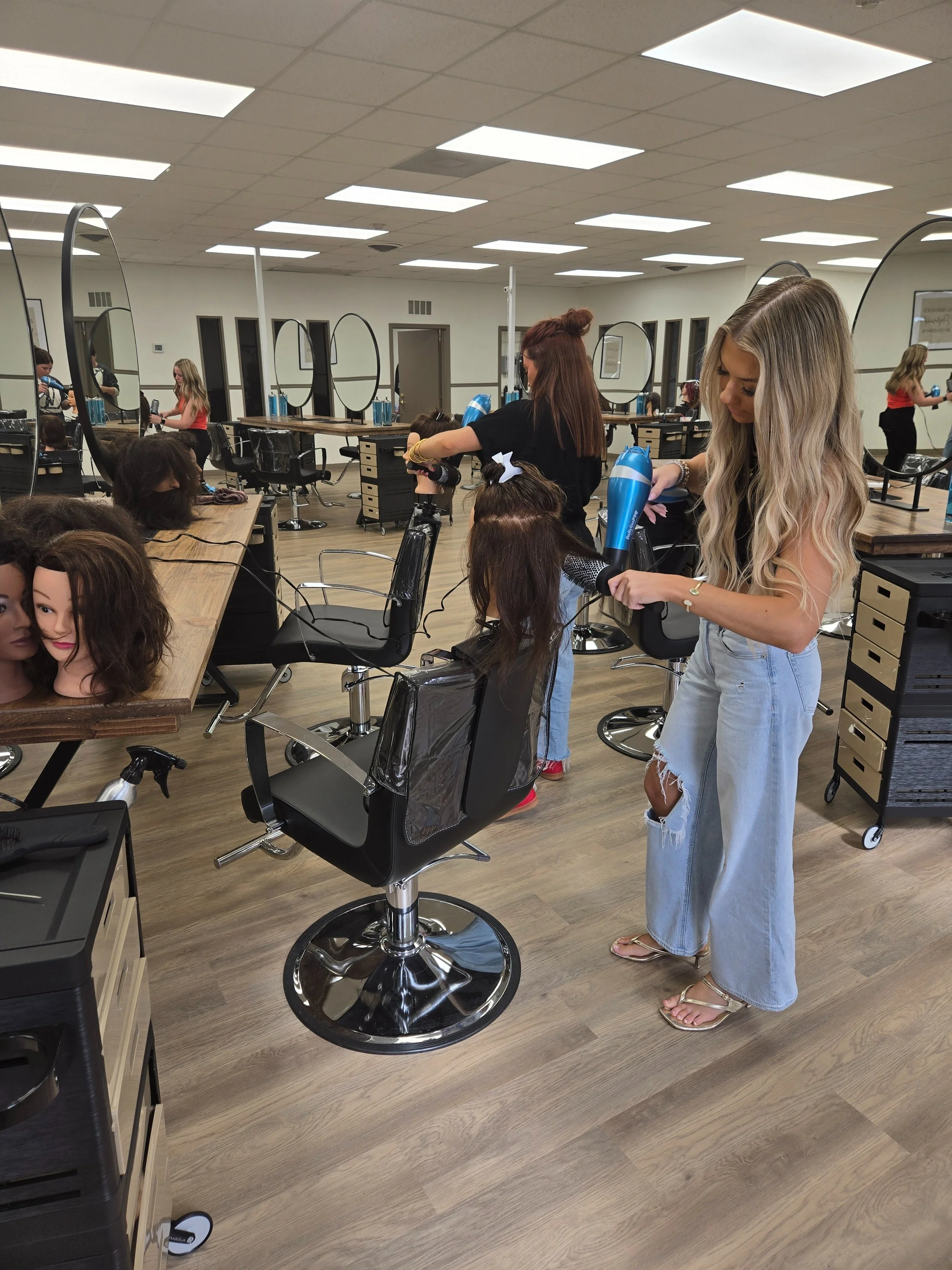 Women working on mannequin heads with hairdryers in a salon, practicing hairstyling.