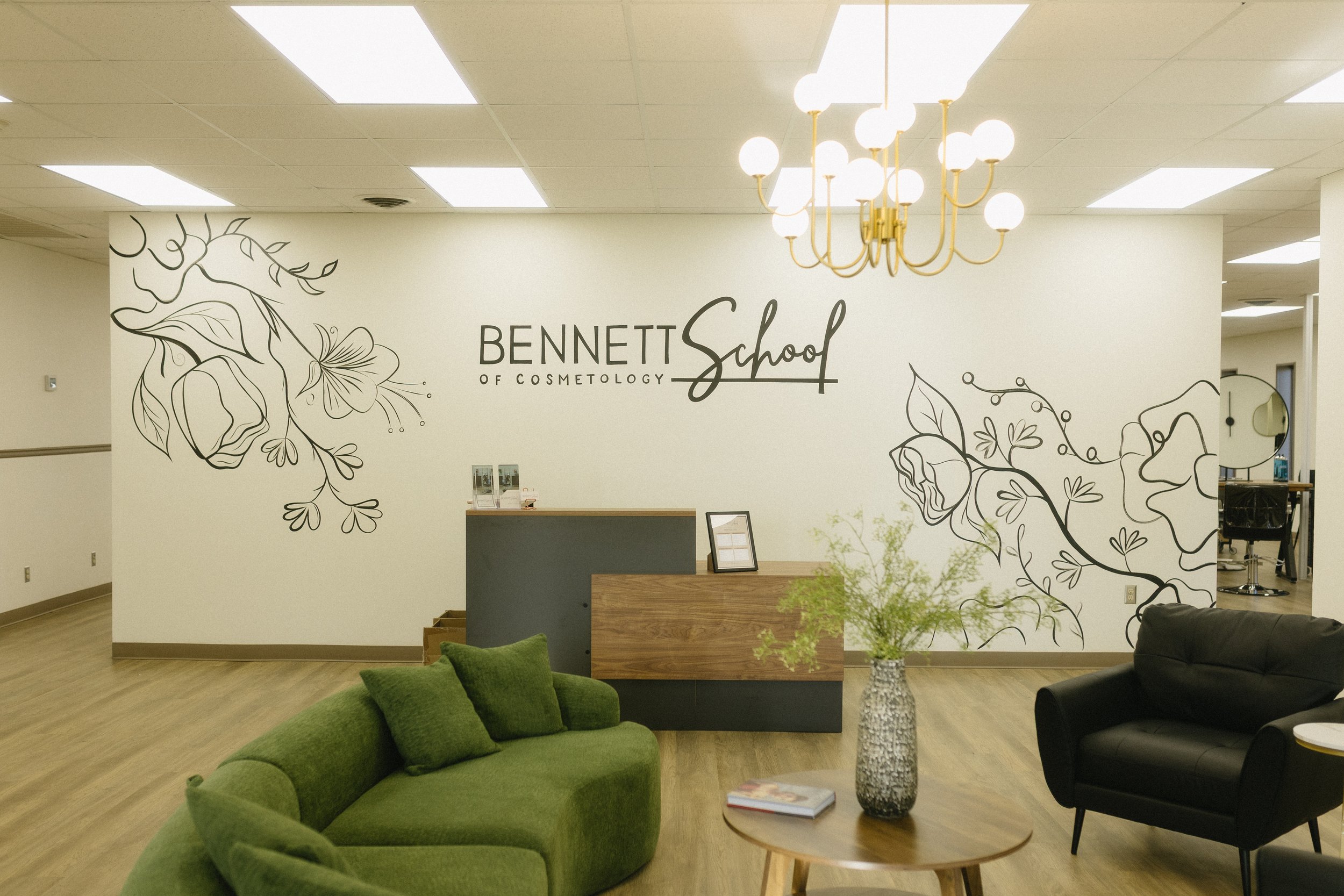 Reception area of Bennett School of Cosmetology with a green sofa, black armchair, round wooden table with a vase of greenery, and decorative wall art of flowers, with a chandelier hanging from the ceiling.