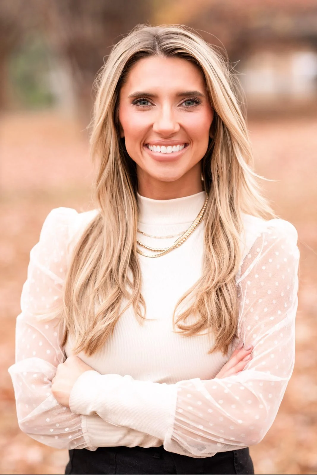 A woman with long blond hair smiling outdoors in fall, wearing a white top with sheer polka-dot sleeves and layered gold necklaces.