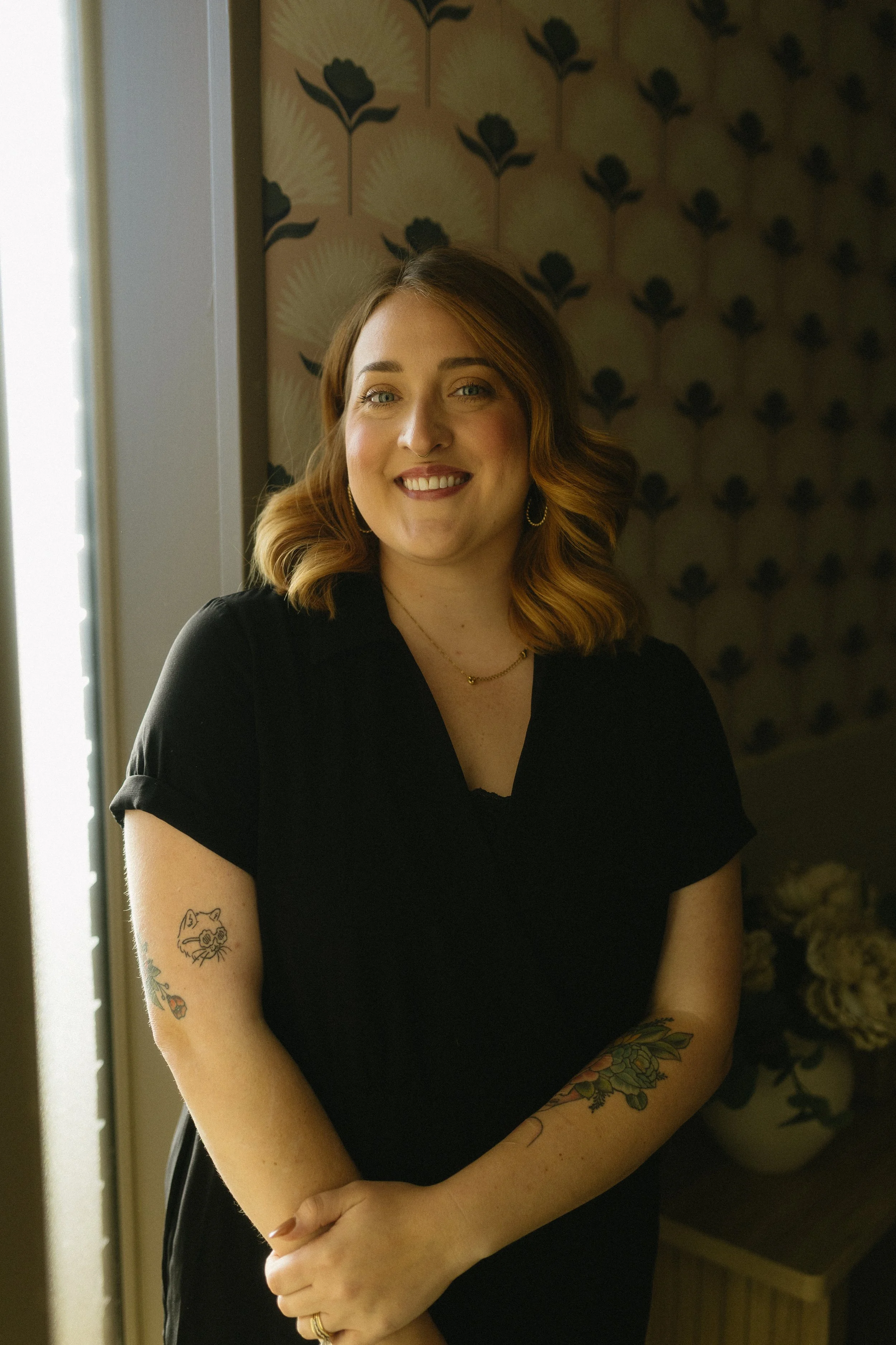 A smiling woman with shoulder-length wavy hair, wearing a black top, standing indoors near a window and a decorative floral wallpaper, with tattoos on her arms.