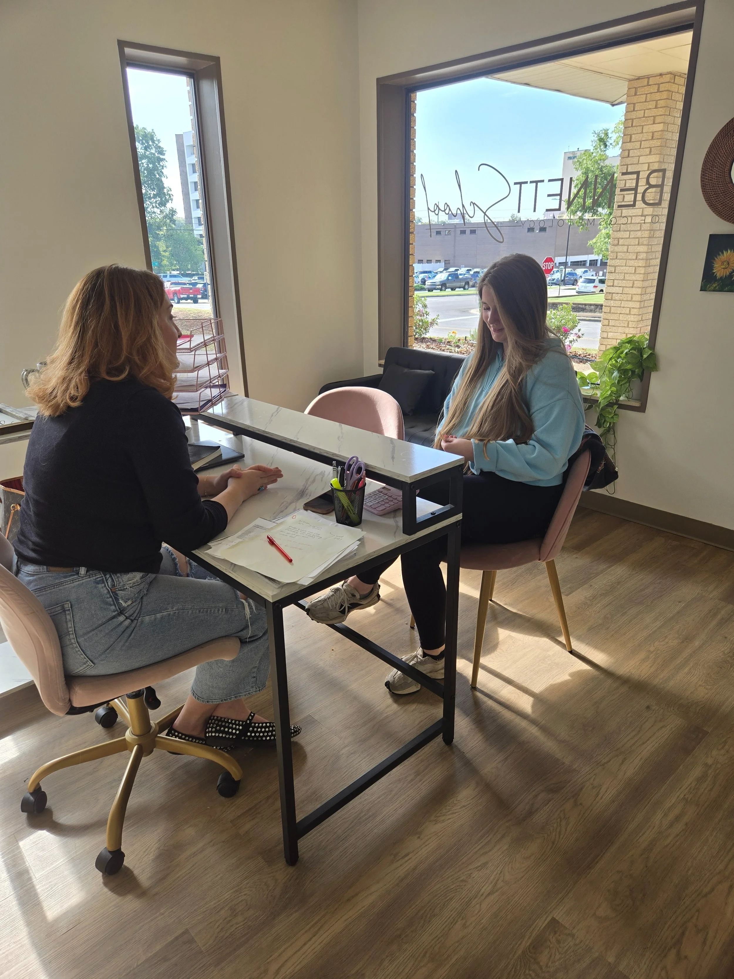 Two women sitting across from each other at a reception desk, having a conversation inside an office with large windows showing a parking lot outside.