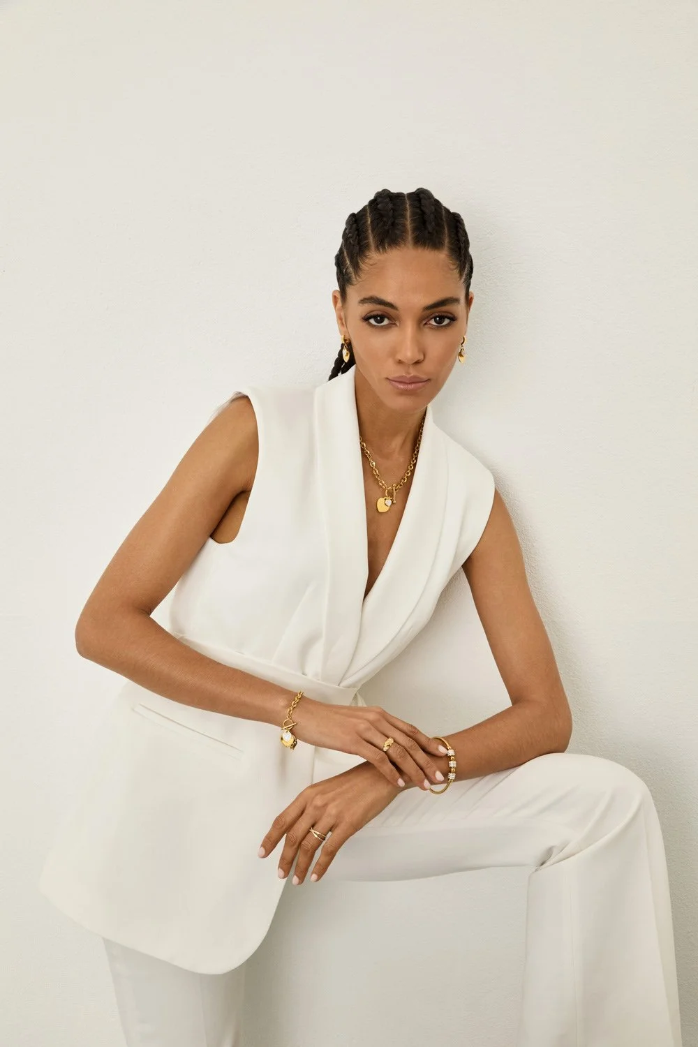 A woman with braided hair and gold jewelry, dressed in a white sleeveless blazer and pants, posing against a plain white wall.