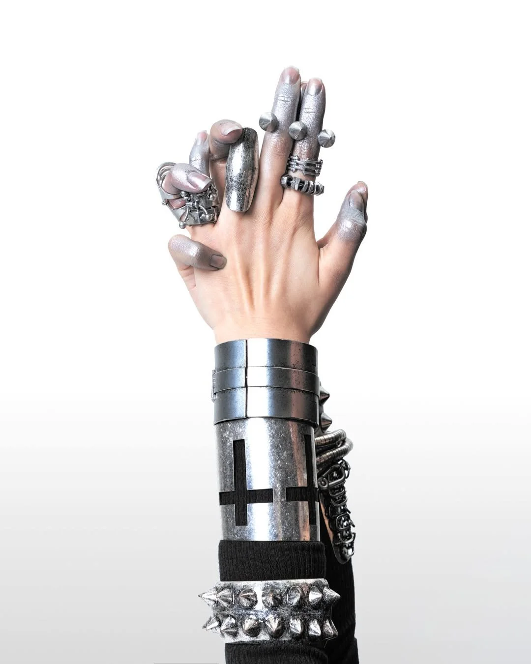 A hand with multiple metallic rings, bracelets, and painted nails, is raised with fingers interlocked, against a white background.