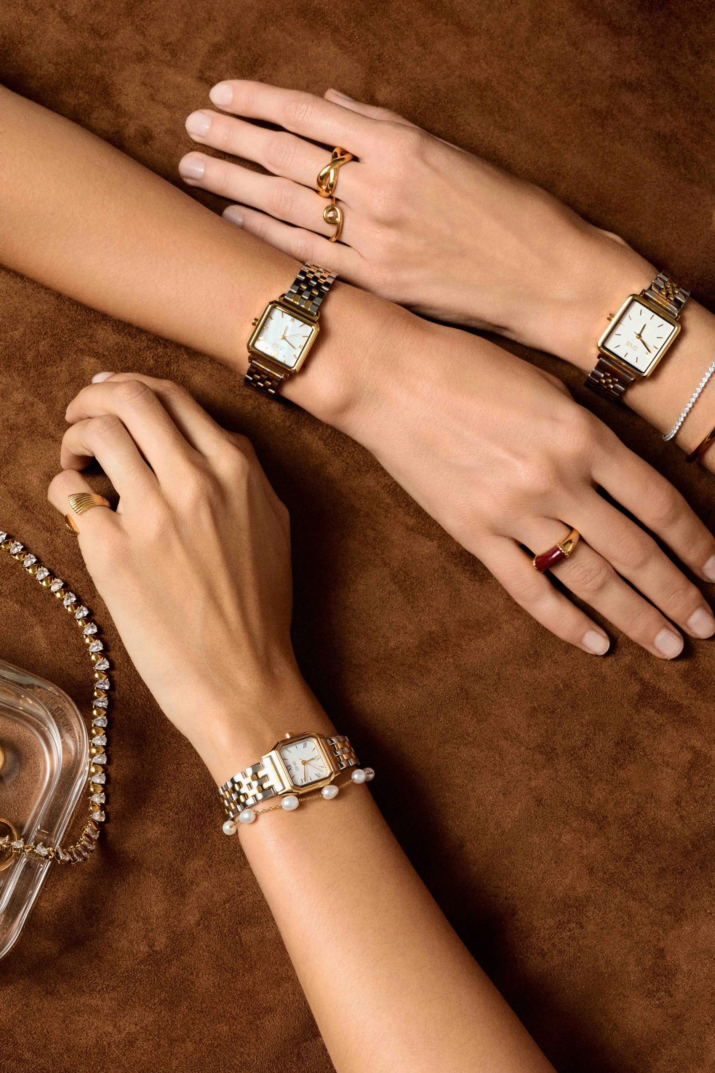 Multiple female hands displaying watches and rings on a brown surface, with jewelry including a pearl bracelet, diamond bracelet, and gold rings.