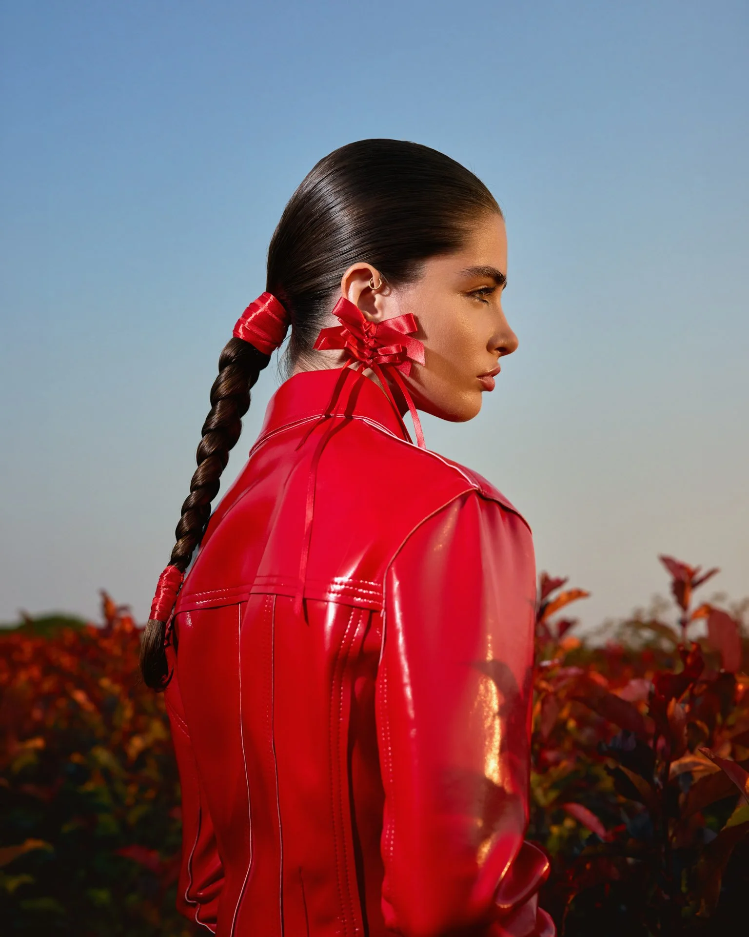 A woman with long dark hair in a braid, wearing a bright red leather jacket and red ribbons in her hair, standing outdoors with a clear sky and reddish-leafed bushes in the background.
