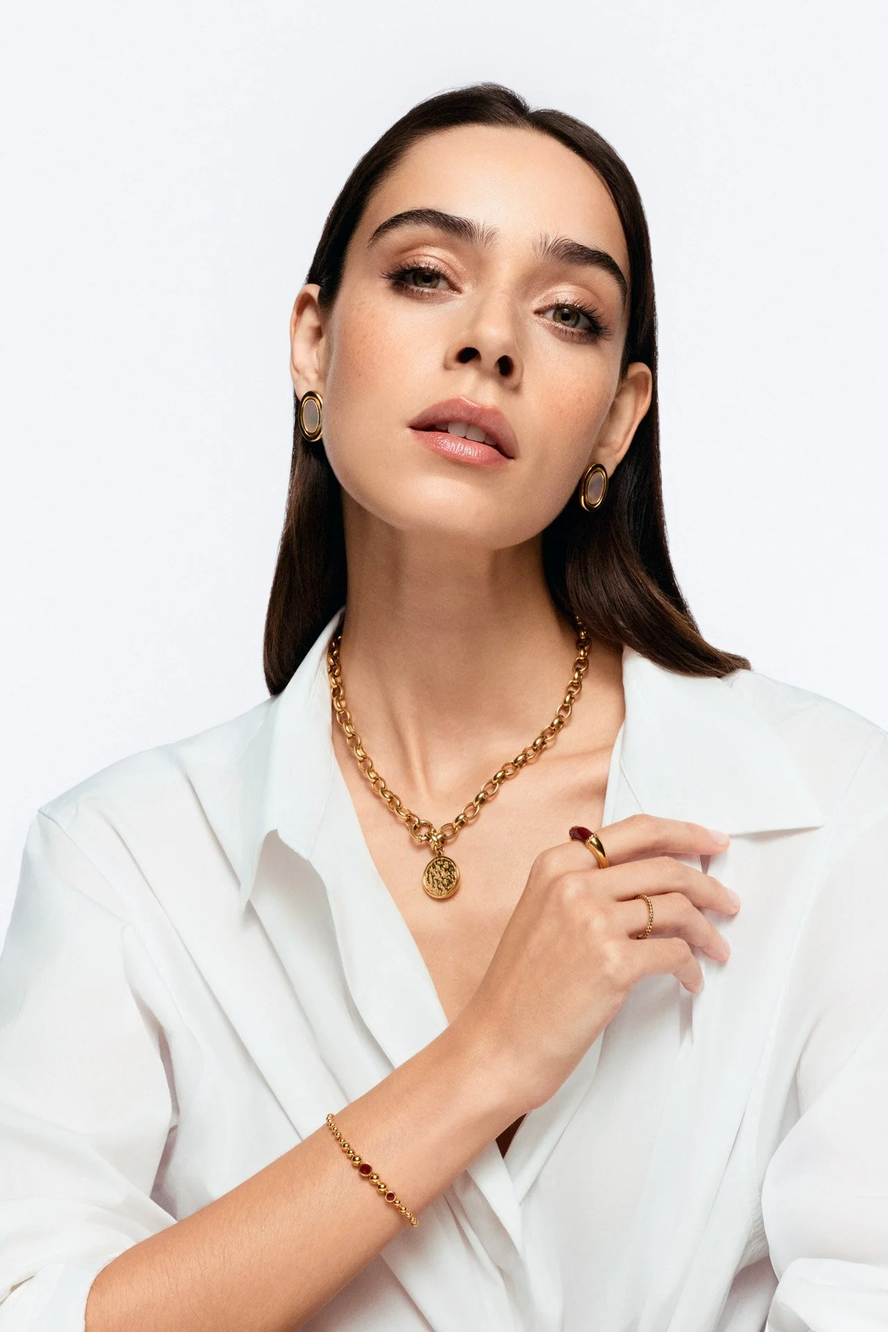 A woman with dark hair wearing gold jewelry, including earrings, a necklace, a bracelet, and rings, dressed in a white shirt against a plain background.