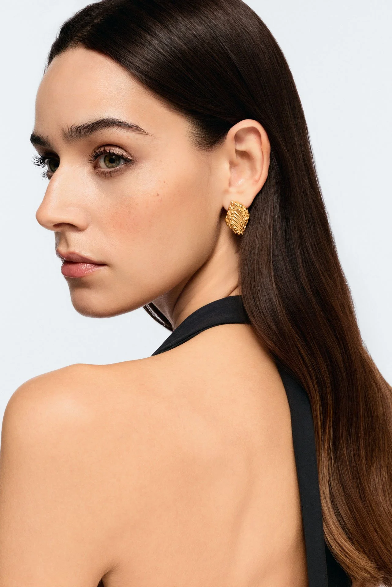 Close-up of a woman with long brown hair wearing gold earrings and a black sleeveless top, looking to the side against a plain background.