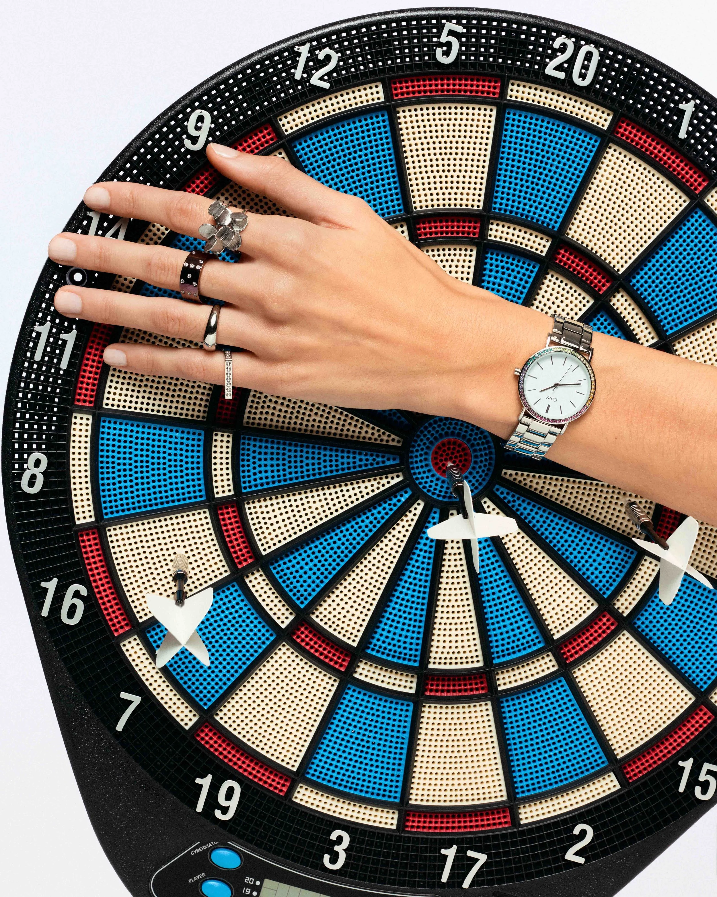 A woman's hand rests on a dartboard, with several rings on her fingers and a silver wristwatch on her wrist.