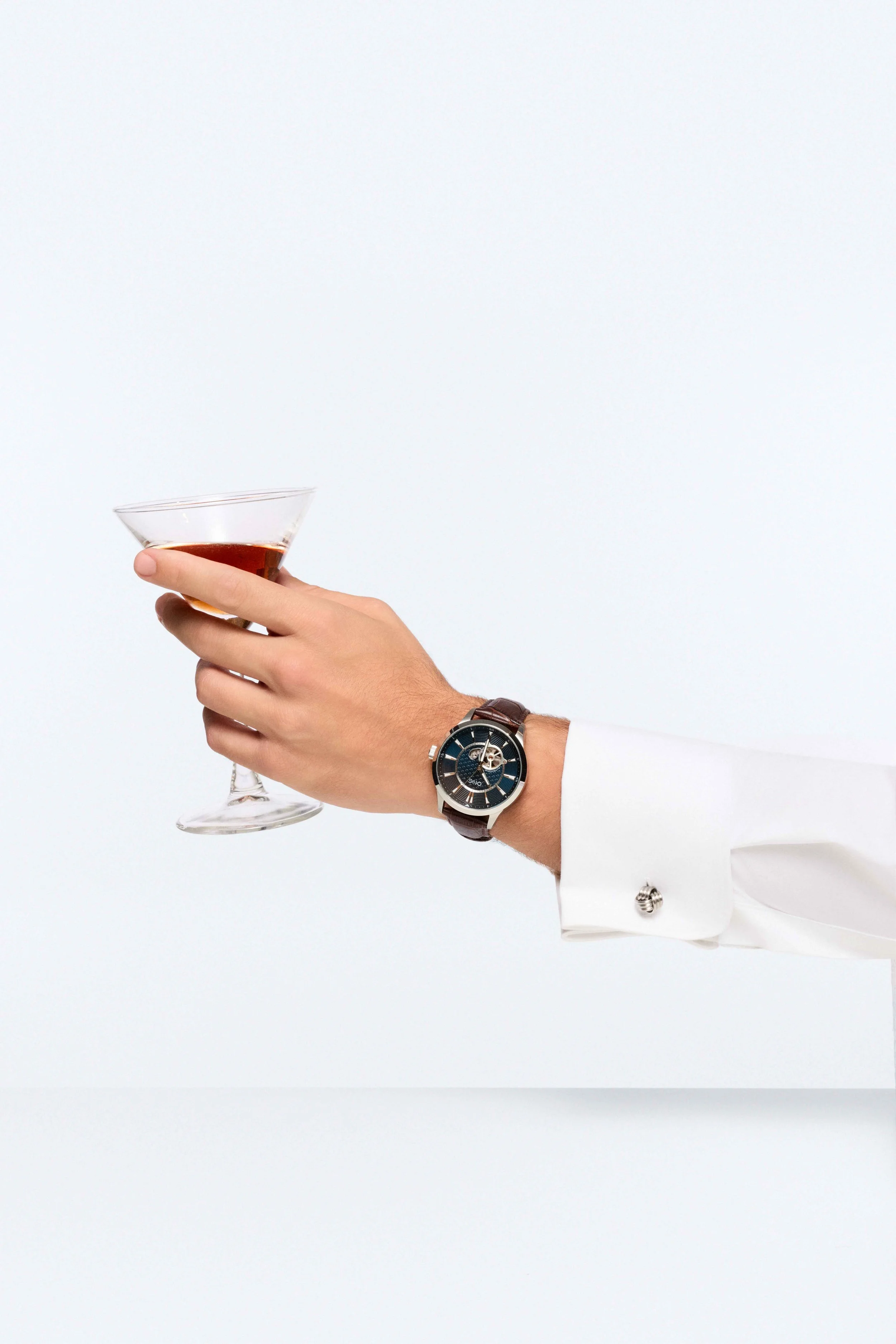 A man's hand holding a martini glass with a brown drink, dressed in a white shirt with a silver cufflink, wearing a large black wristwatch with a leather strap.