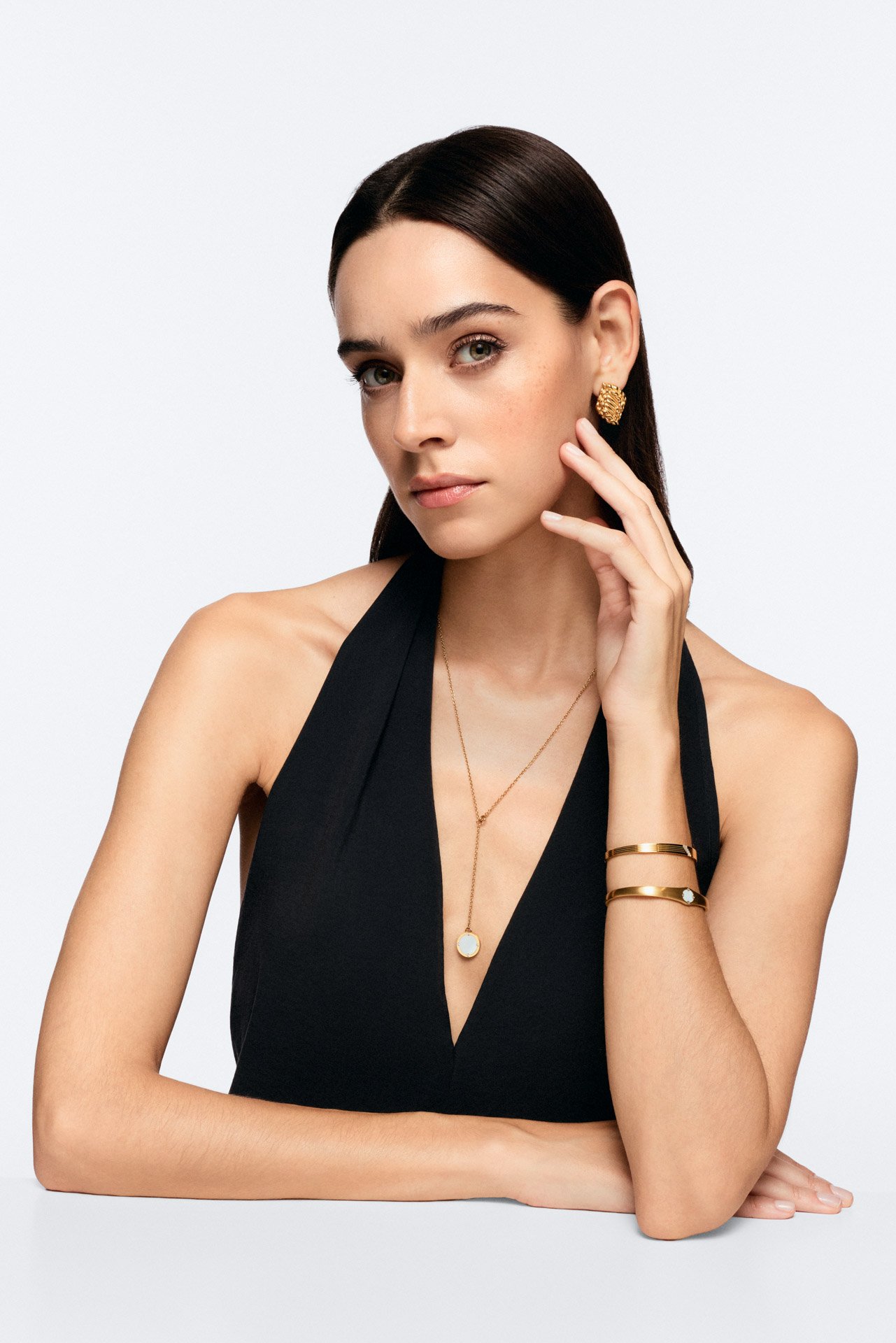 A woman with long dark hair, wearing a black sleeveless top, gold jewelry including earrings, bracelets, and necklaces, posing against a plain white background.