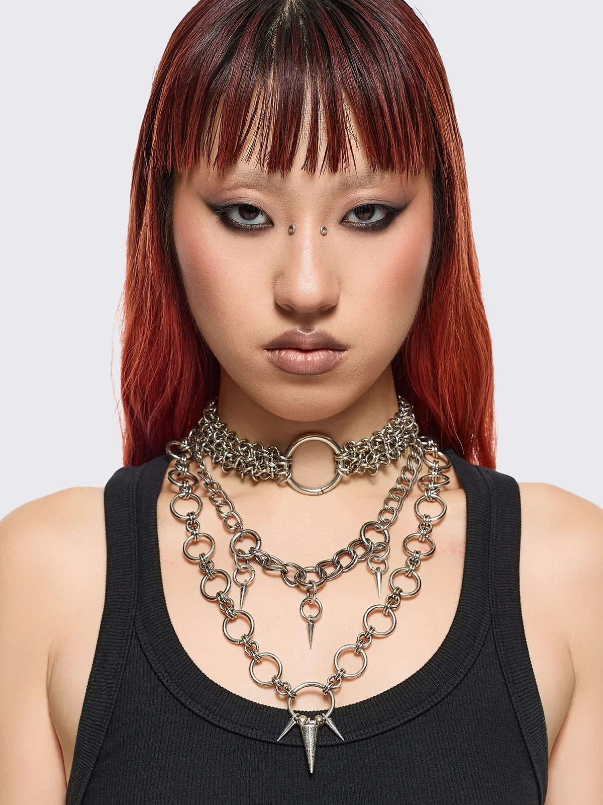 A woman with red hair and makeup wearing multiple layered silver chains with spikes and rings, and a black tank top.