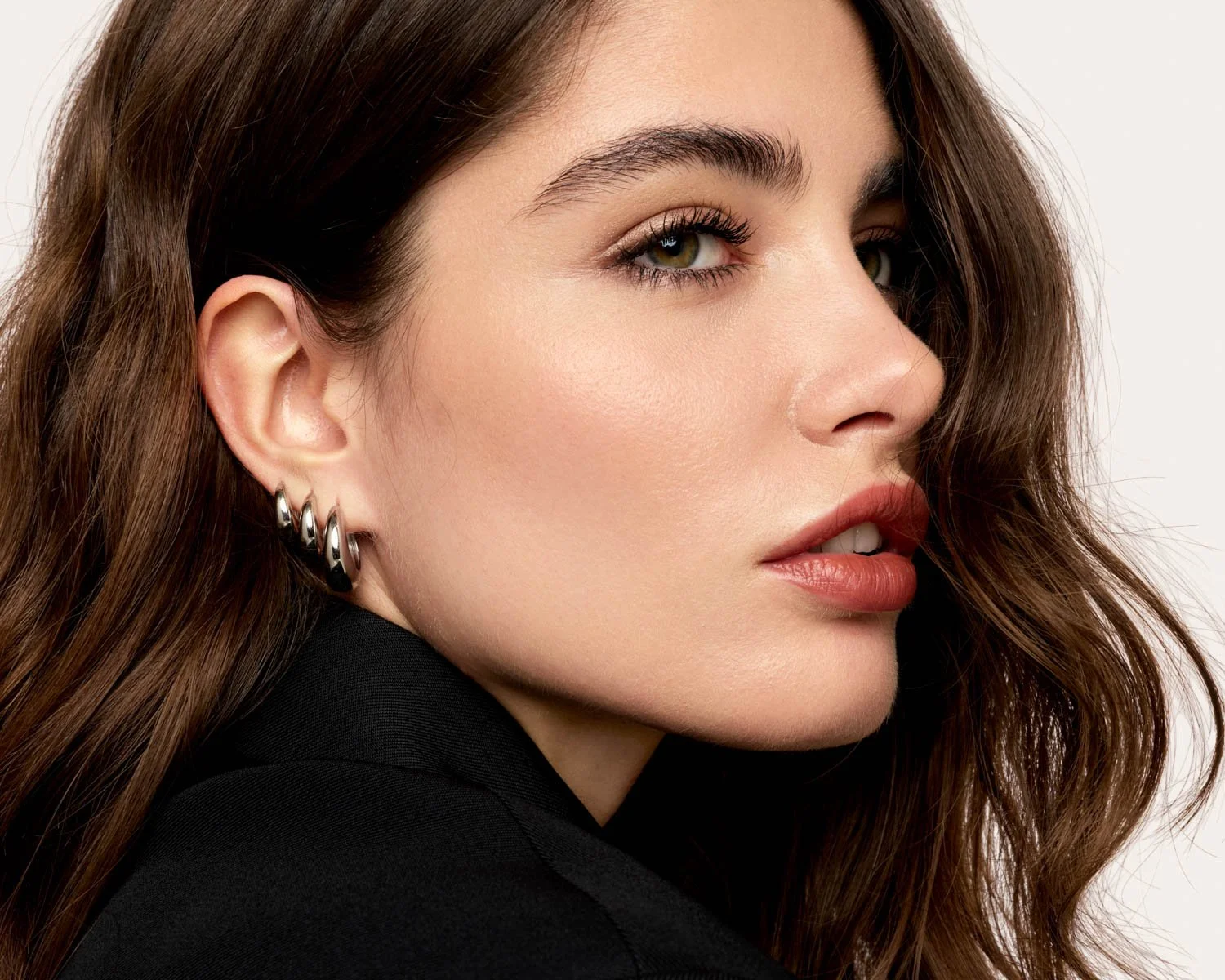 A close-up of a woman's face showing her with long brown hair, hoop earrings, light makeup, and a black top, looking slightly to the side against a plain background.