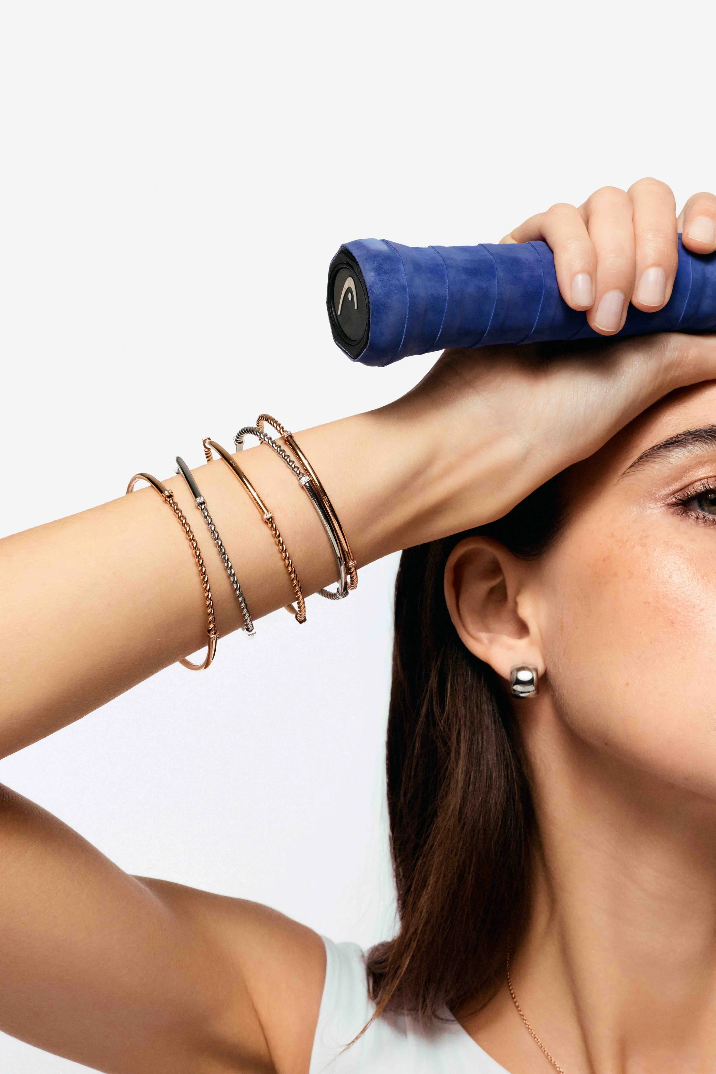 A woman with brown hair, wearing earrings and bracelets, holds a blue tennis racket against her forehead, with part of her face visible.
