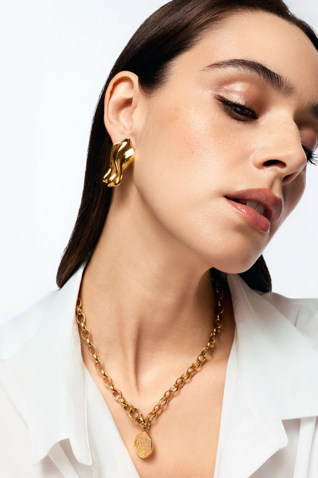 Close-up of a woman wearing large gold earrings and a matching gold chain necklace, with a white shirt and natural makeup.
