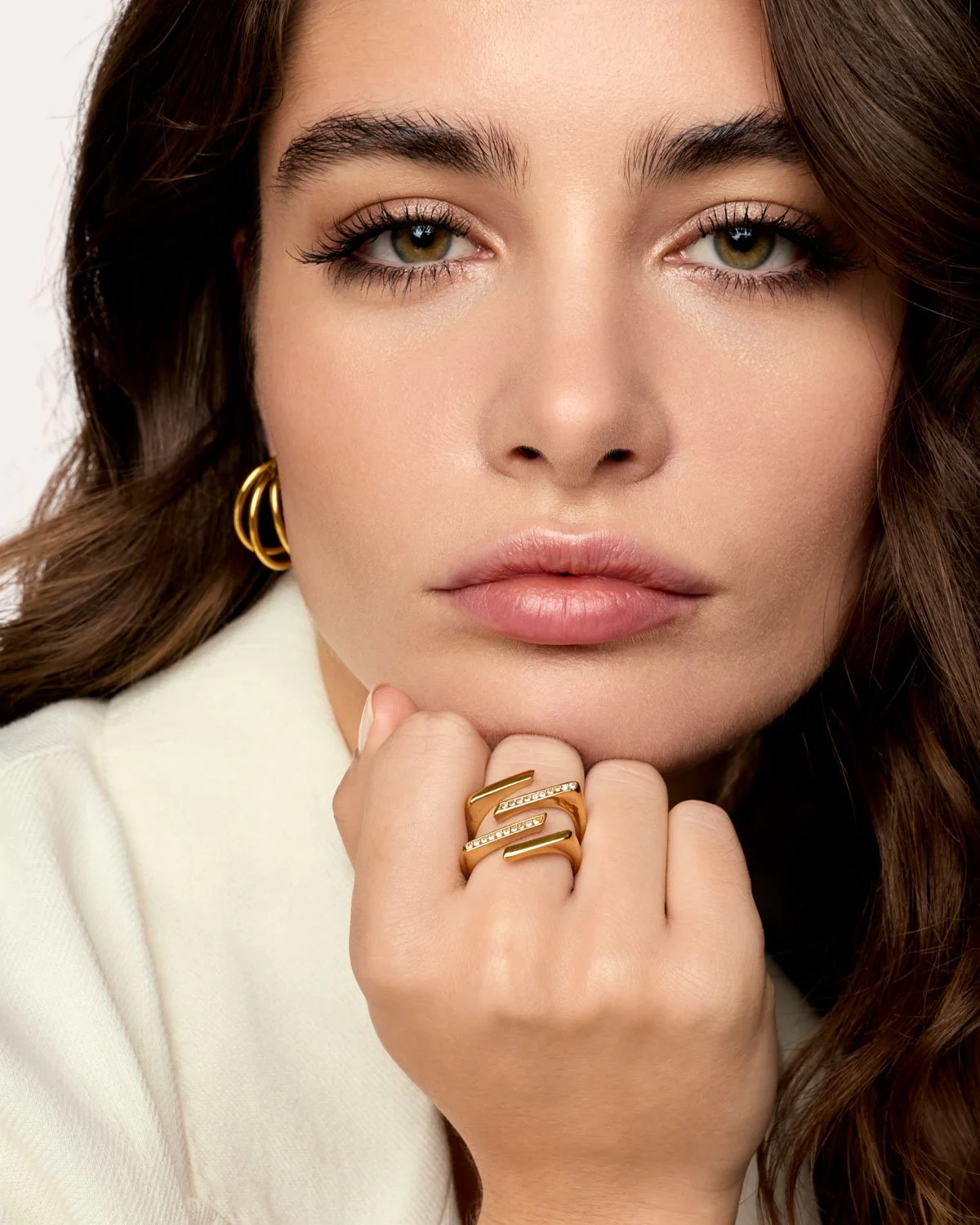 Close-up of a woman with fair skin, hazel eyes, and brunette hair, wearing gold hoop earrings and a gold ring with a zigzag design and embedded diamonds, resting her chin on her hand.