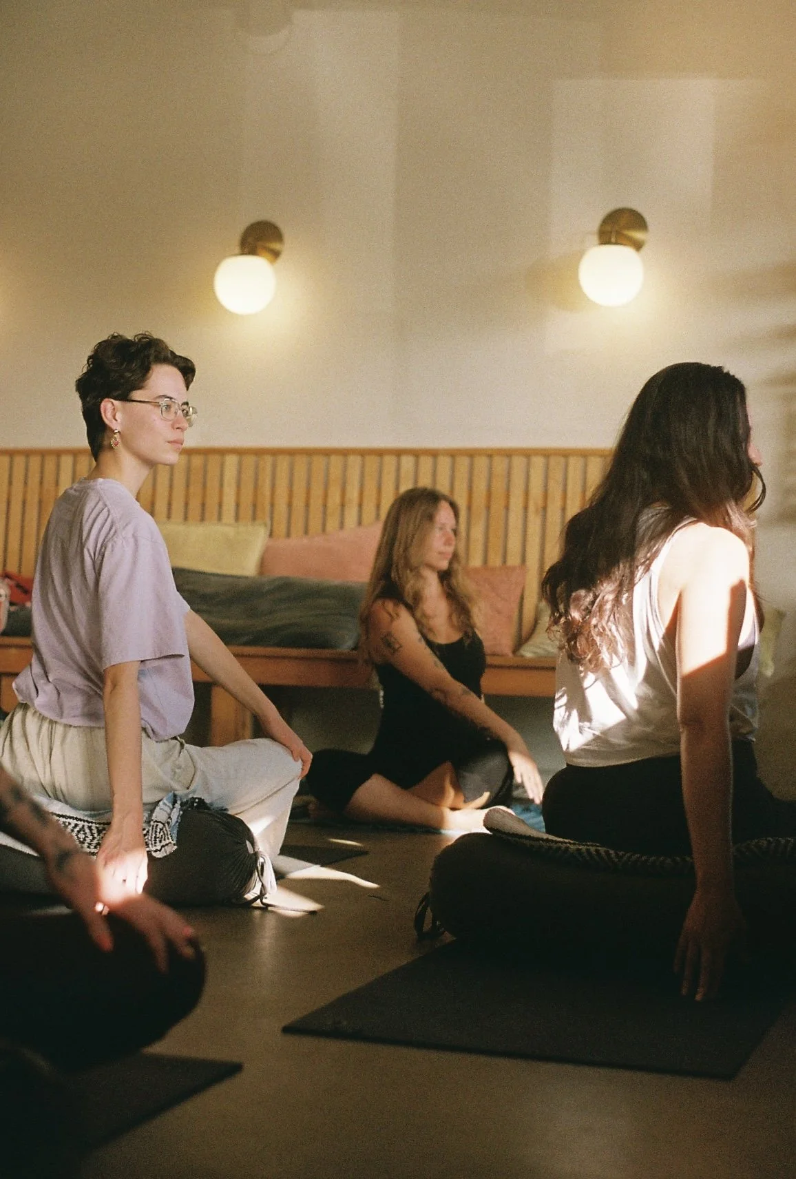 Yoga teacher sitting at the front of the room, guiding students in a seated spinal twist looking over shoulder.