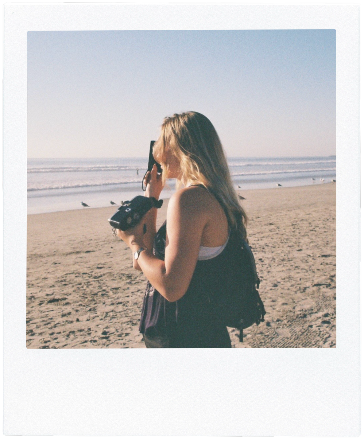 35mm film photo of Sarah Madore taking a photo with a film camera on the beach. Framed with a white border that resembles an instant film photo. Link to @saltedgranola TikTok page.