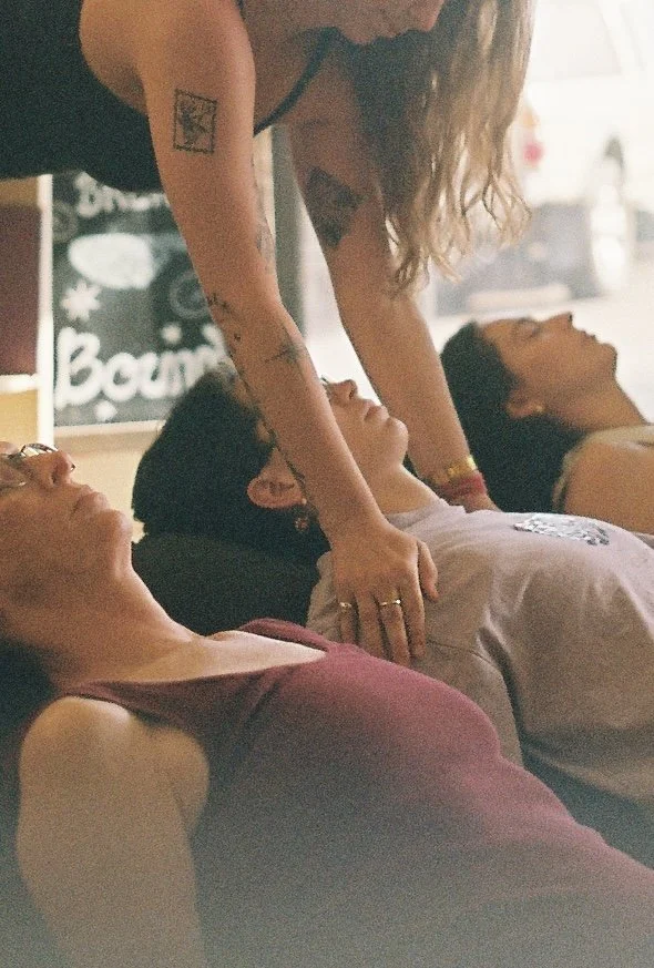 Close up of yoga students laying face-up on bolsters while yoga teacher provides hands-on adjustments