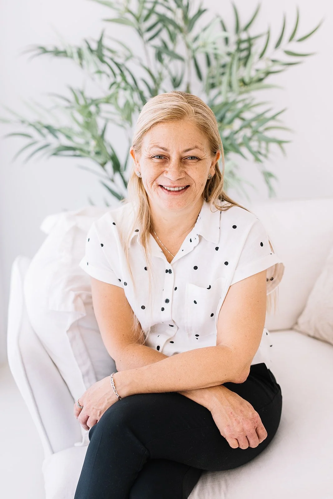 A middle-aged woman with long blond hair, wearing a white blouse with black polka dots and black pants, sitting on a white couch with a green plant in the background.
