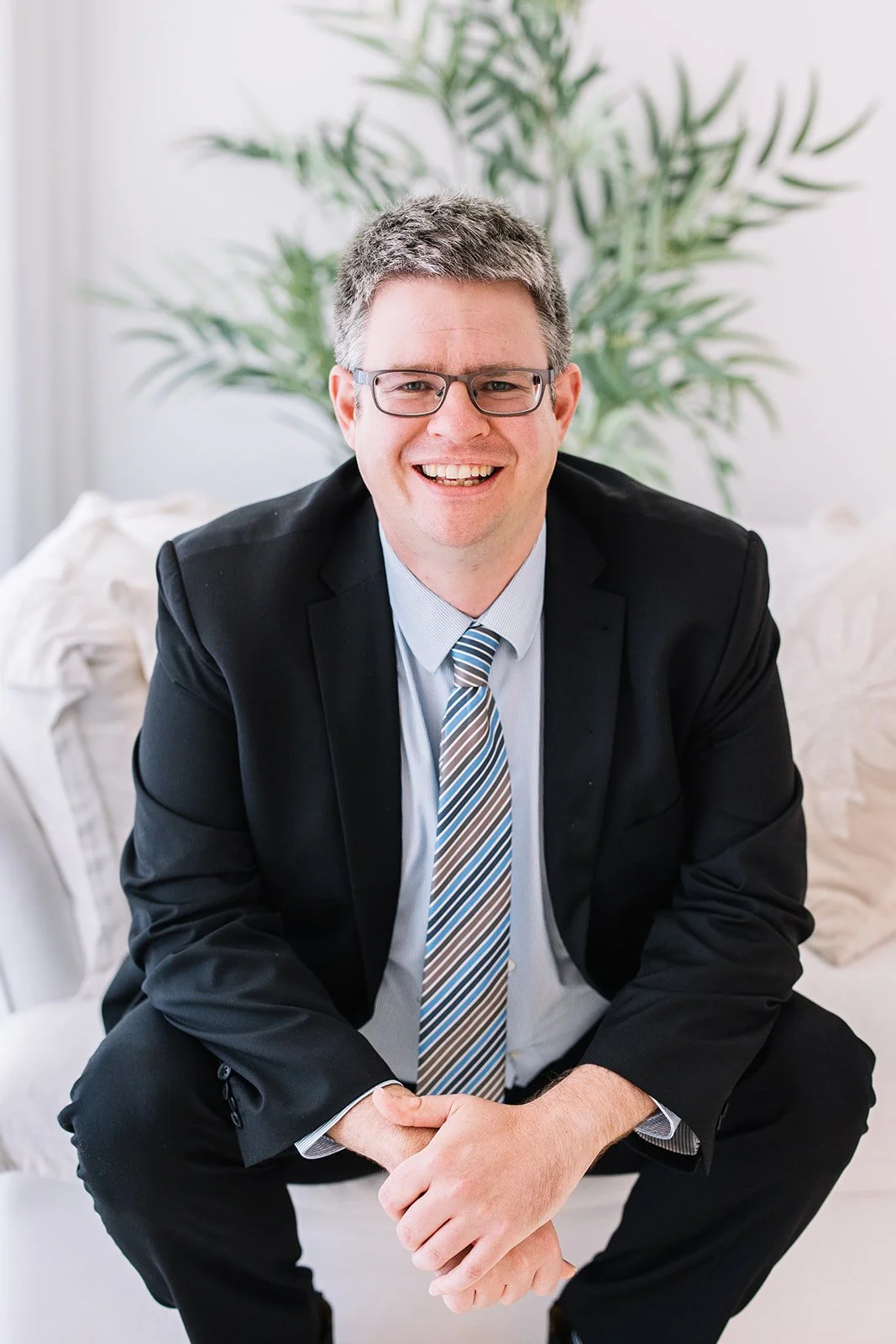 Man in black suit and tie sitting on a white couch in front of a large green plant, smiling