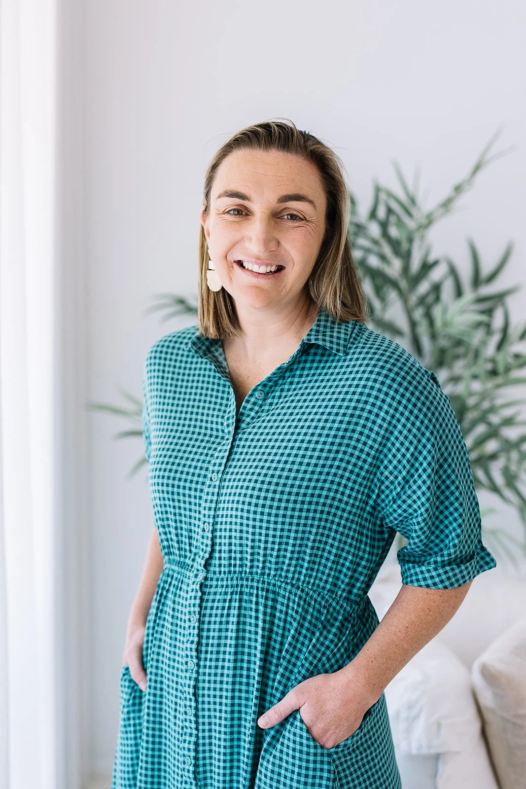 A smiling woman with shoulder-length brown hair wearing a teal and black checkered dress standing indoors in front of a white wall and green plant.
