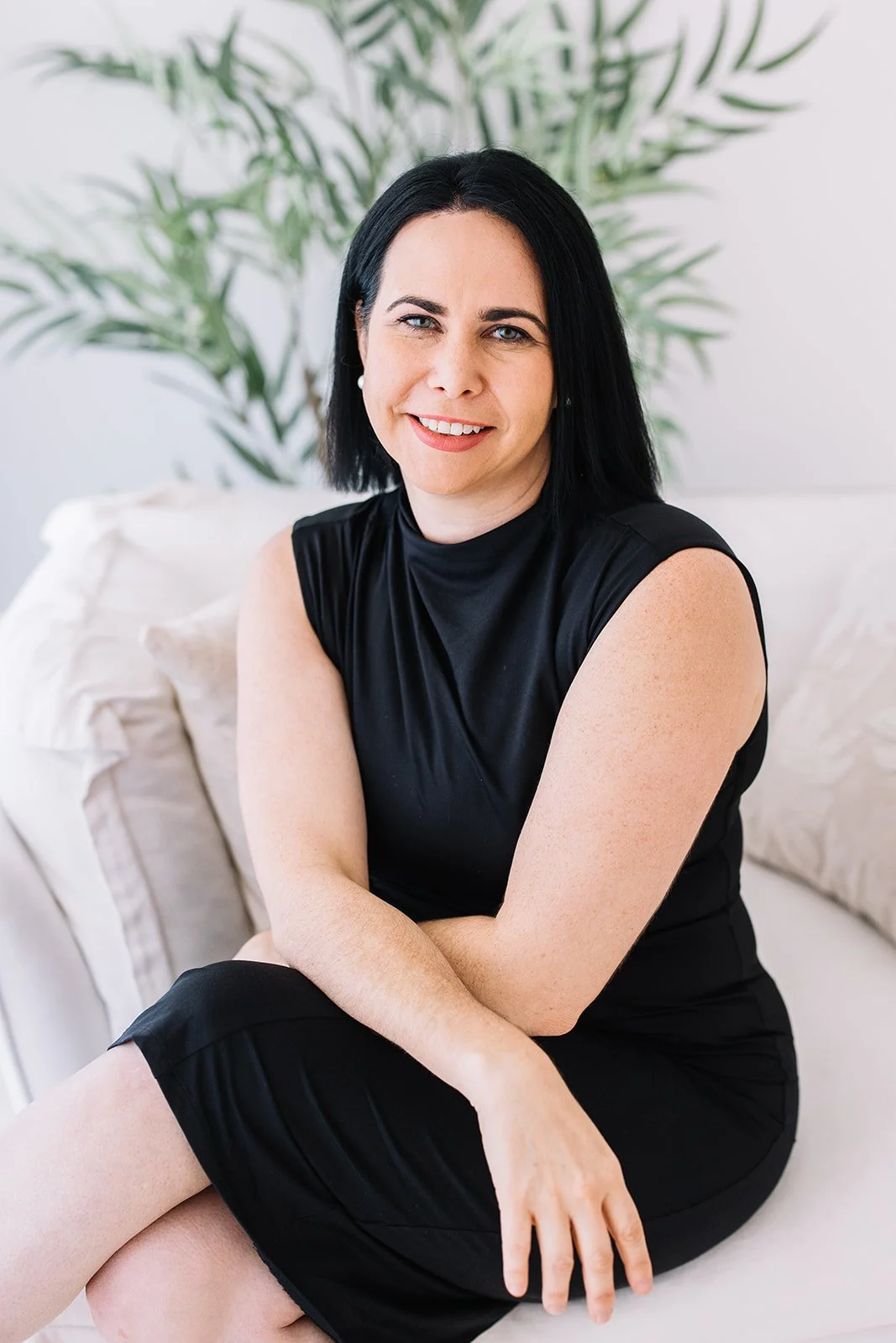 A woman with black hair and blue eyes sitting on a white sofa, wearing a black sleeveless dress, with a large green leafy plant in the background.