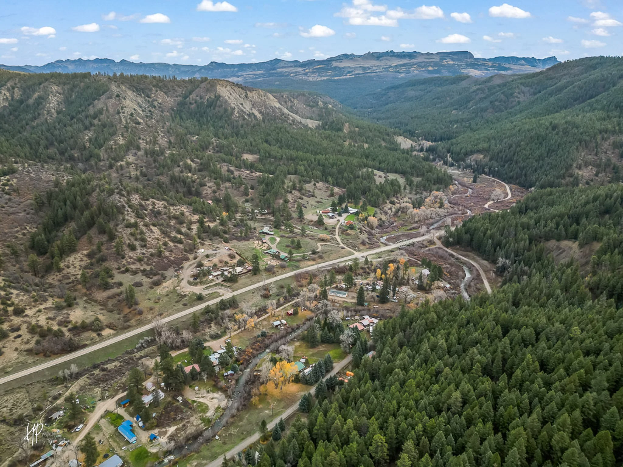Aerial view of a mountainous landscape with a small residential area, winding roads, lush green forests, and distant mountain ranges under a partly cloudy sky.