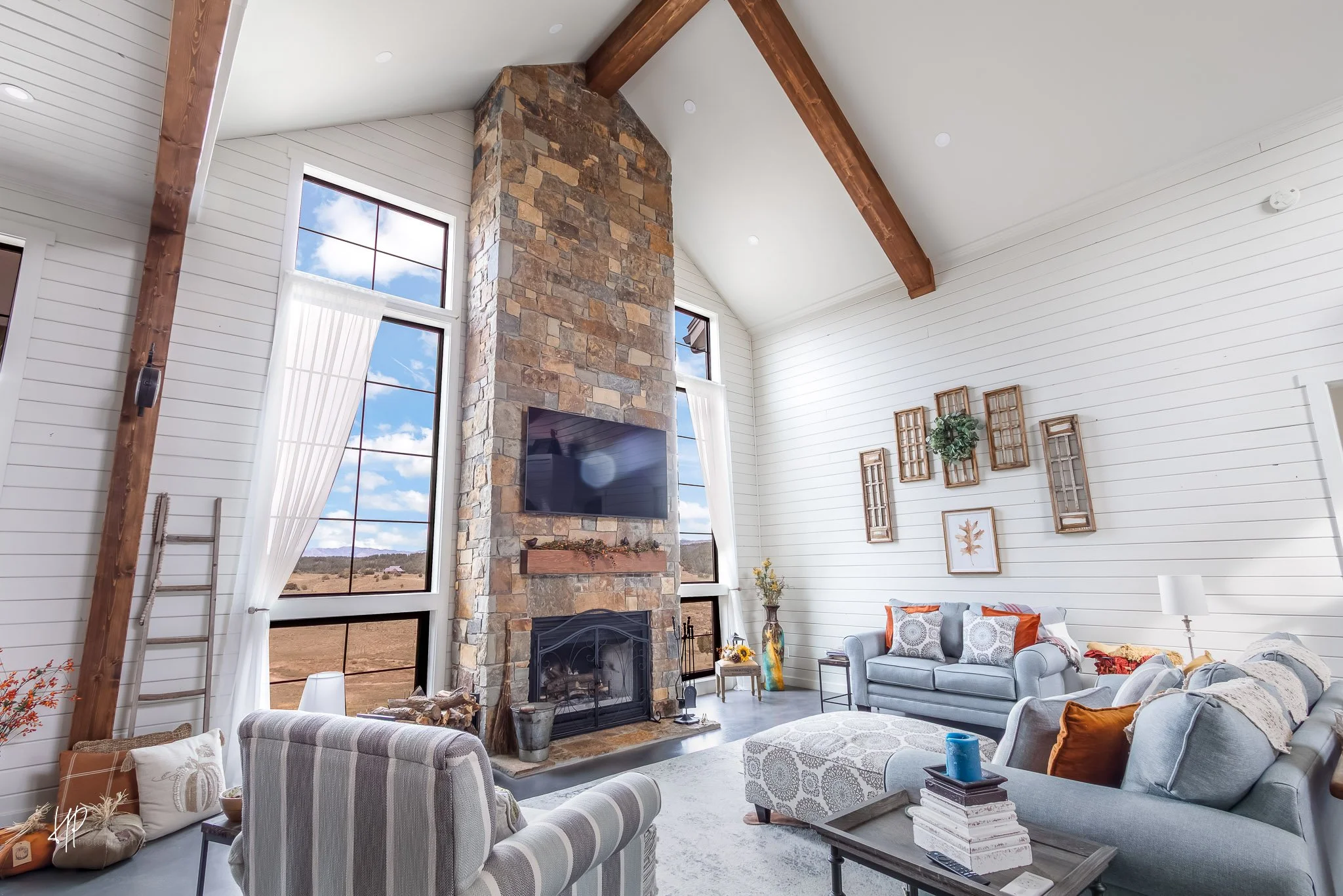 Living room with high vaulted ceiling, stone fireplace, large windows with white curtains, white shiplap walls, and light blue furniture including sofas and armchairs, decorated with pillows and wall art.
