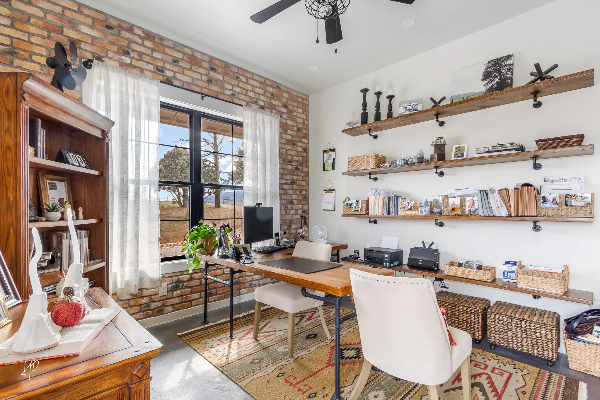 Home office with brick wall, large window, wooden desk, white chairs, shelves with books and decor, computer, and office equipment.