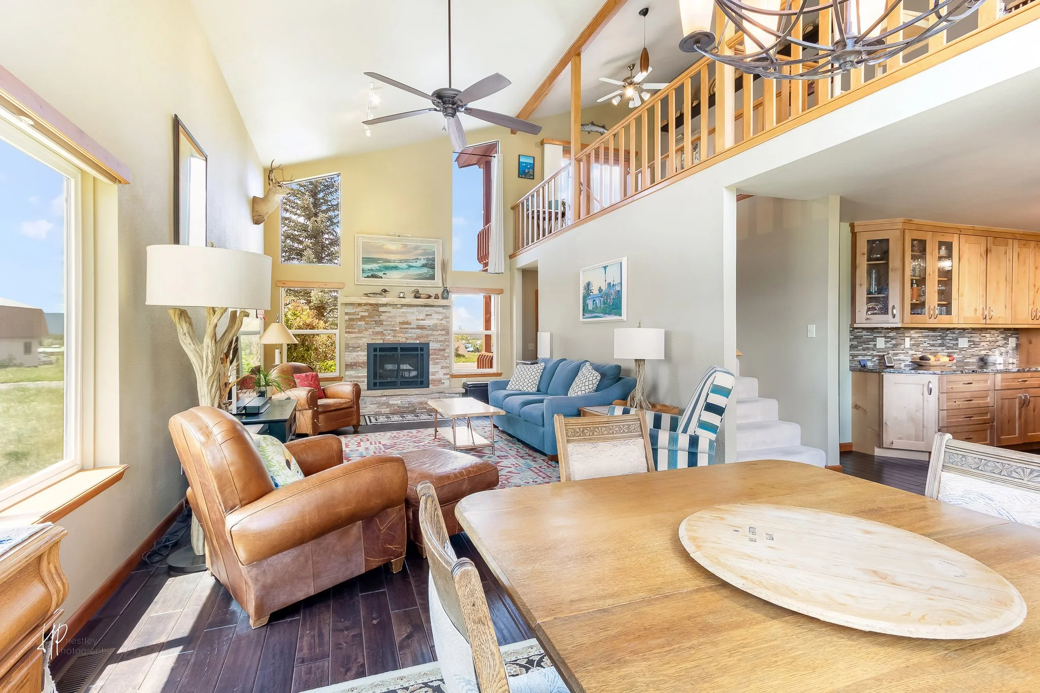 Living room with high vaulted ceiling, large windows, a fireplace, and mixed furniture including leather chairs, a blue sofa, and a dining table.