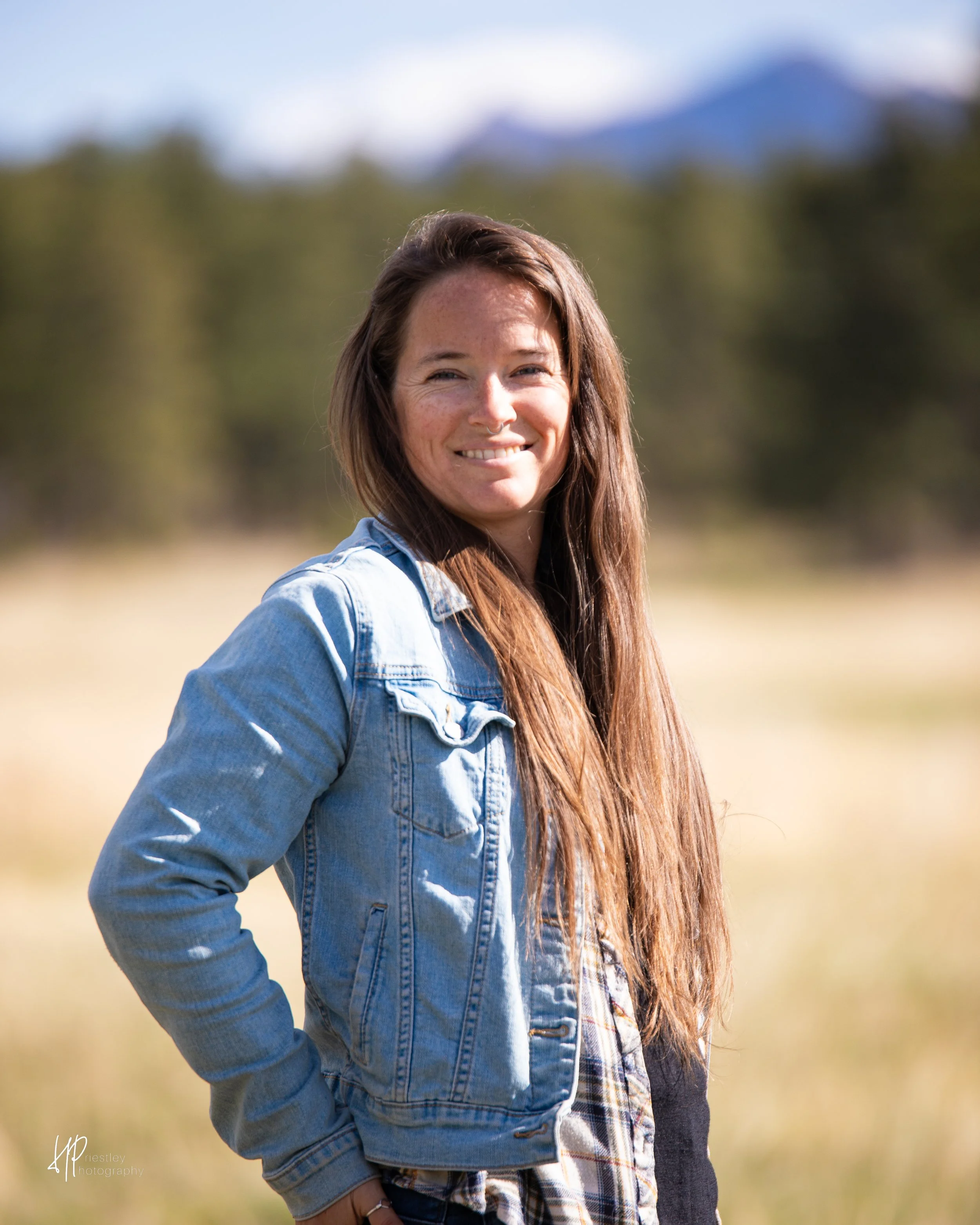 A woman with long brown hair wearing a light blue denim jacket and plaid shirt standing outdoors with a blurred background of trees and a mountain in the distance.