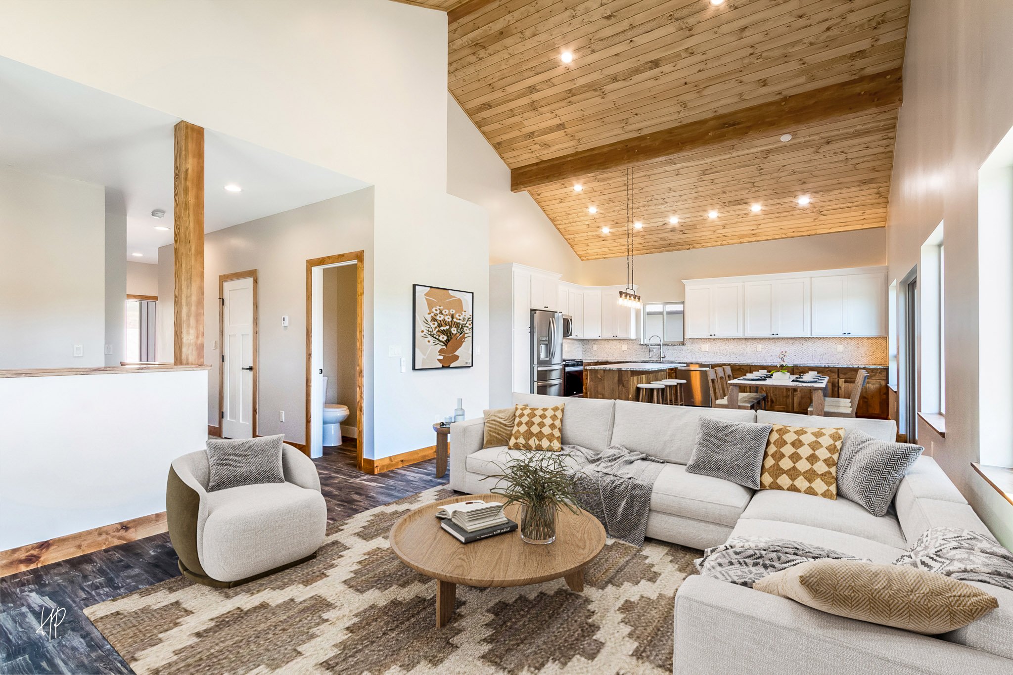 Open-concept living room and kitchen with a white sectional sofa, beige and patterned pillows, and a round wooden coffee table with books and a plant. The room has a high, wood-paneled vaulted ceiling with recessed lighting. The kitchen features white cabinets, stainless steel appliances, and a wooden island with barstools. A small artwork hangs on the wall near a doorway leading to a bathroom.