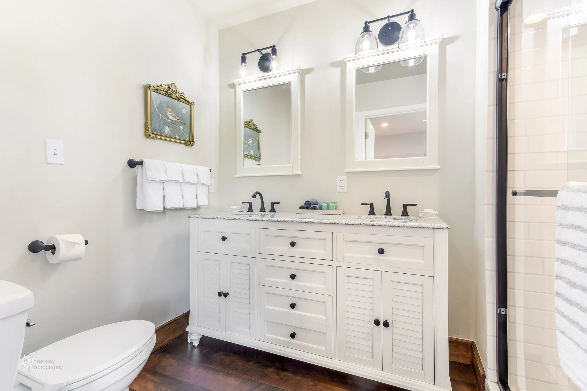 Bathroom with white vanity, two mirrors, black faucets, towel, toilet paper, art, wood floor, shower with glass door.