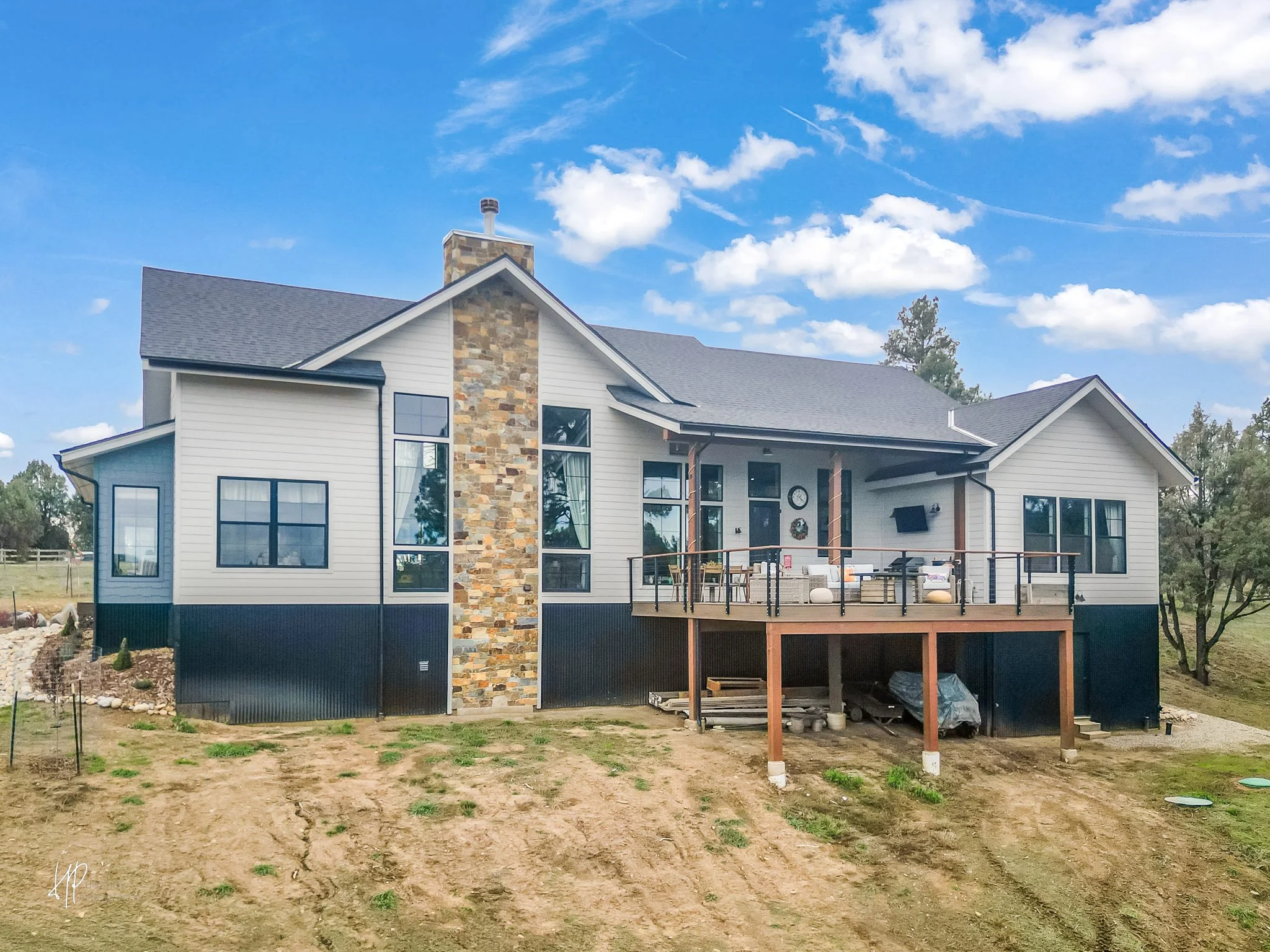 Newly constructed two-story house with a large deck, stone chimney, and multiple large windows, surrounded by a grassy yard and blue sky with clouds.