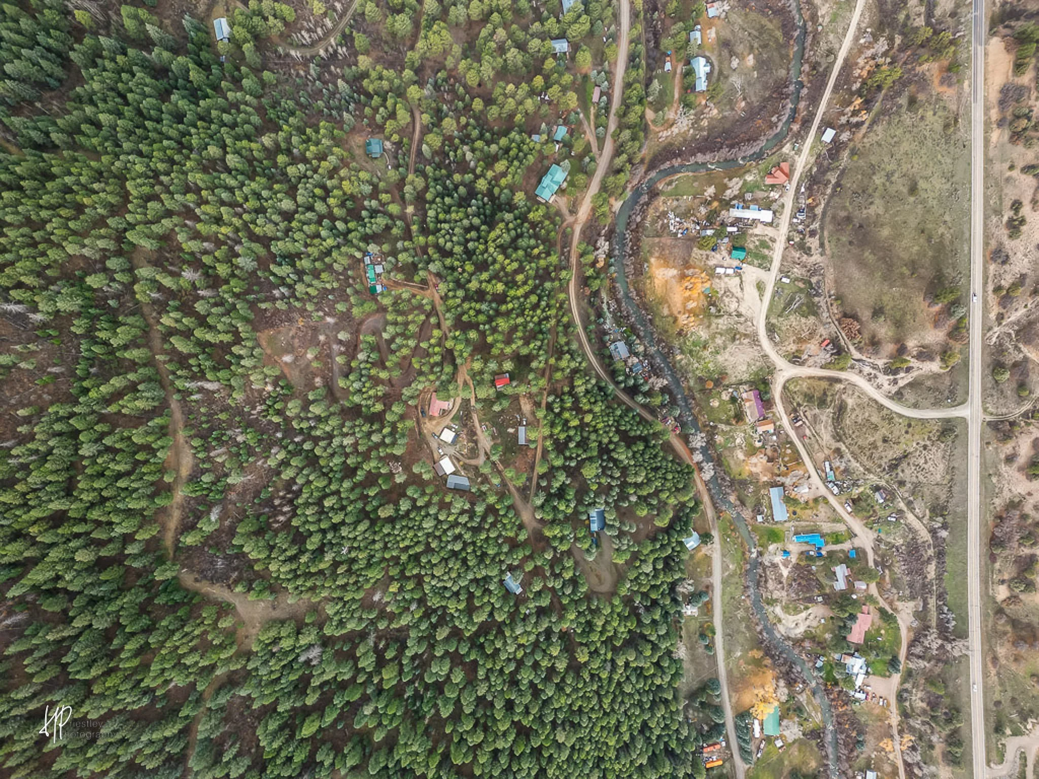 Aerial view of a rural area with dense forest on the left and a small residential area on the right, separated by a winding creek and dirt roads.