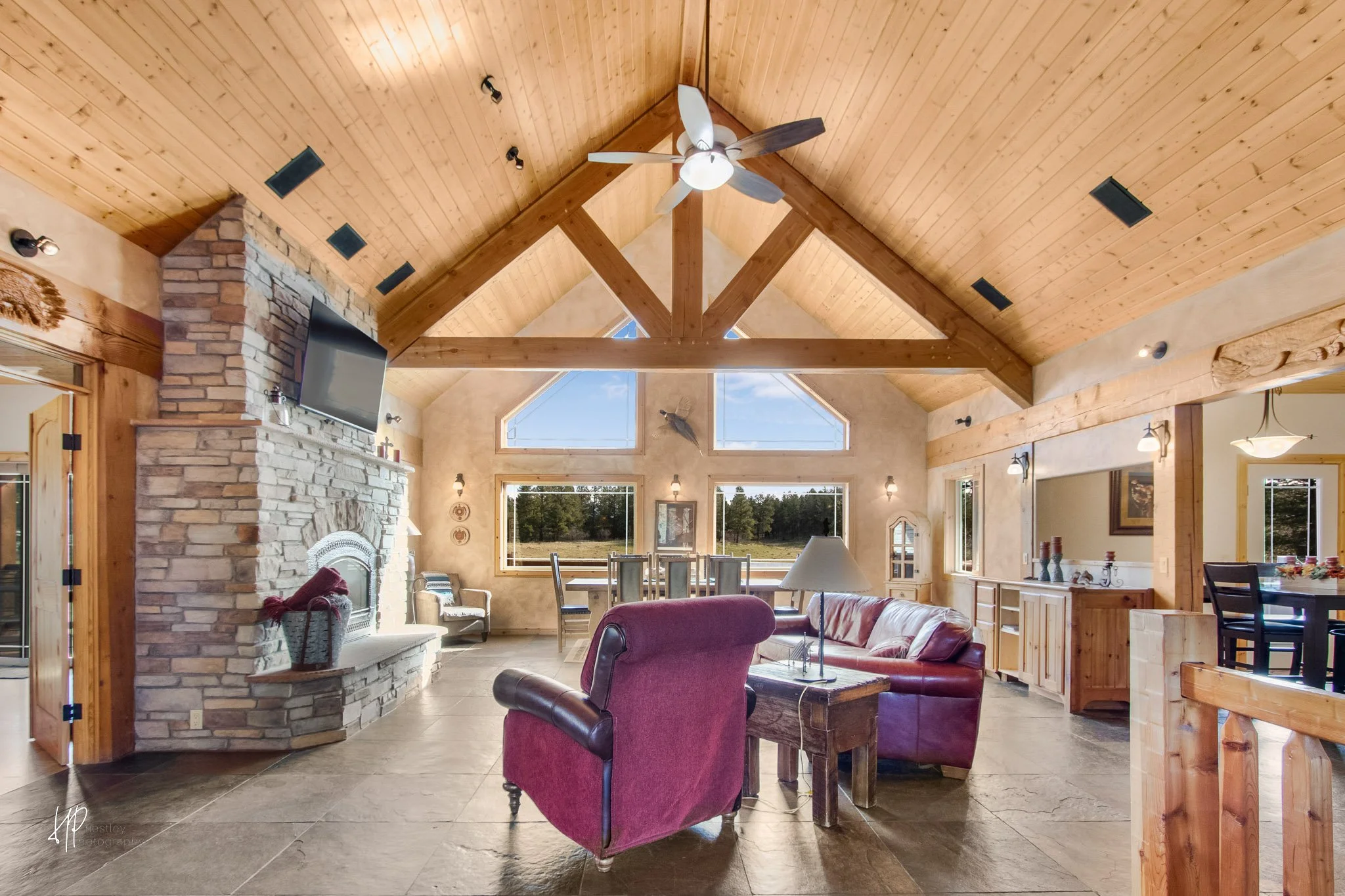 Spacious living room with high wooden ceiling, stone fireplace, large windows showing outdoor landscape, red armchair, matching sofa, dining area, and decorative wall accents.