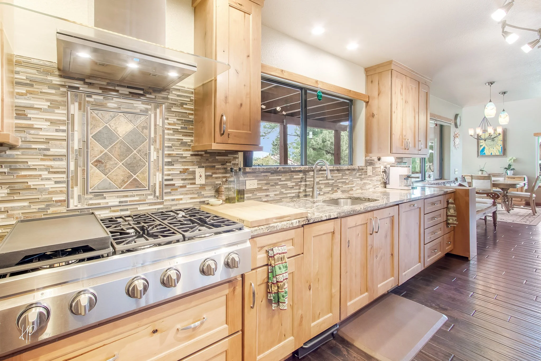 Kitchen with wooden cabinets, a gas stove, a mosaic tile backsplash, a window over the sink, and a dining area in the background.