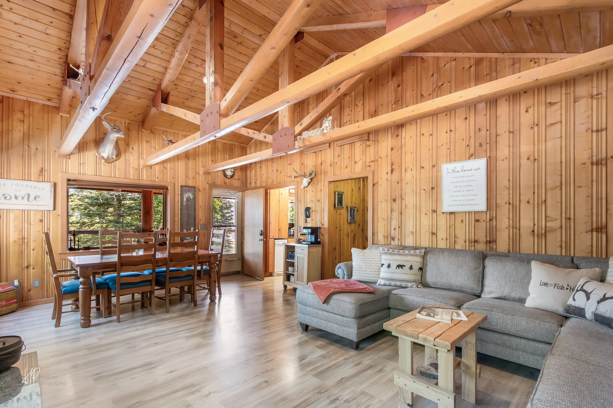 Interior of a wood-paneled cabin living room with a vaulted ceiling, deer and bobcat mounted on the walls, a dining table with chairs, a gray sectional sofa, decor signs, and a view of trees outside through windows.
