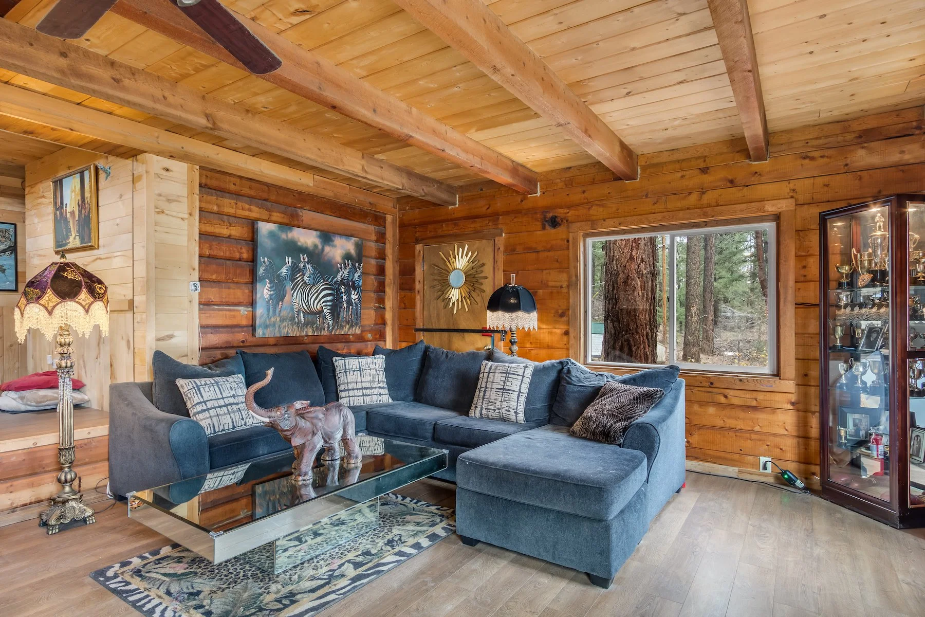 Living room with wooden walls and ceiling, blue sectional sofa with patterned pillows, glass coffee table with an elephant figurine, framed zebra art, window showing trees outside, display cabinet with trophies and pictures.