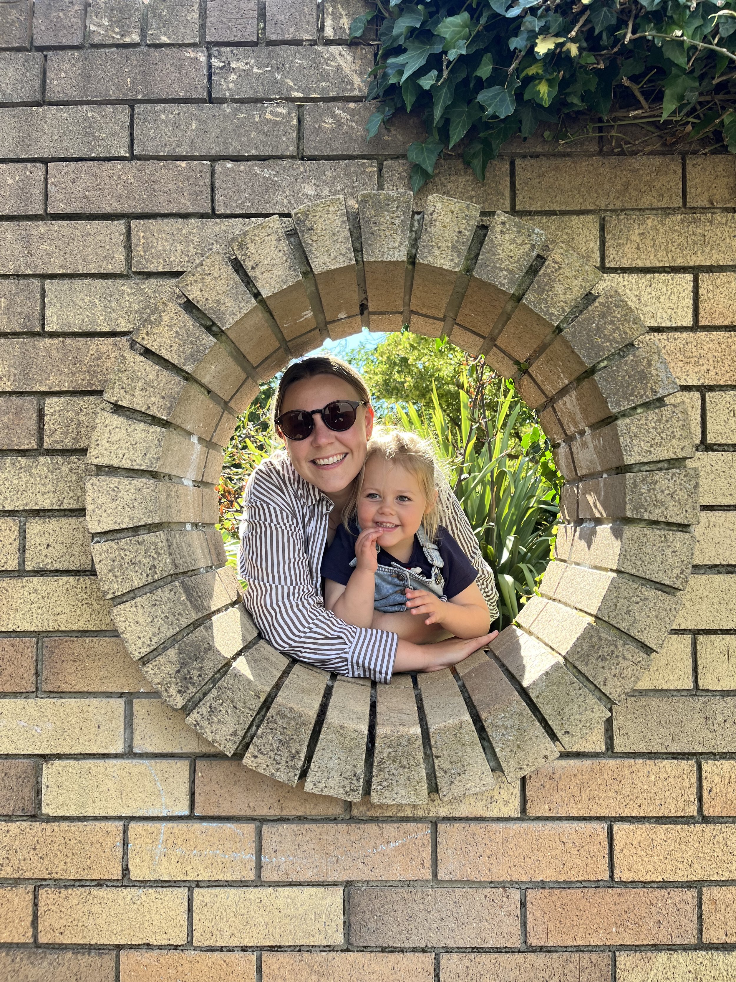 A woman and a young girl are smiling and posing together through a small circular opening in a brick wall, with greenery in the background.