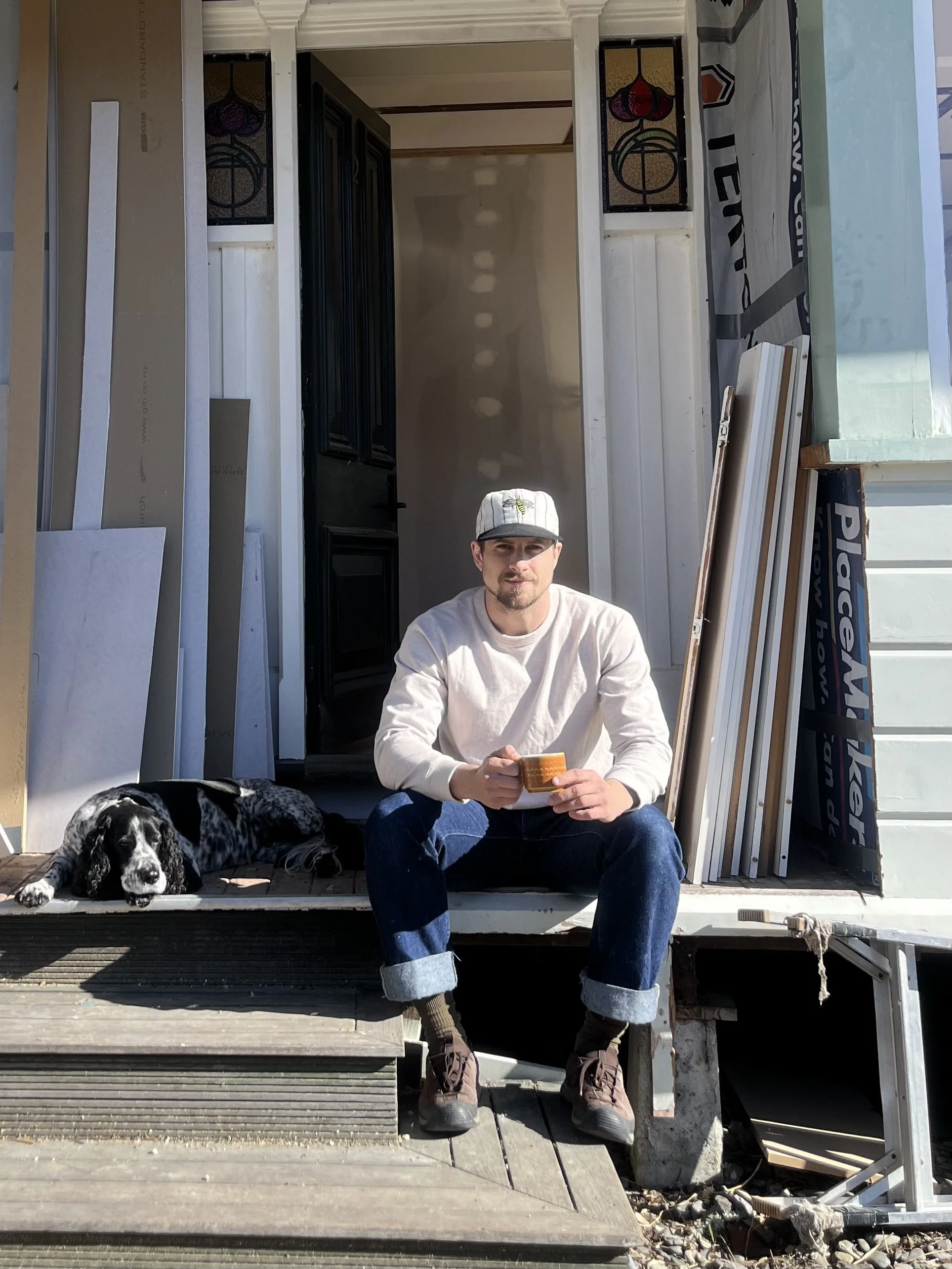 A man sitting on the porch of a house under construction, holding a cup, with a black and white puppy lying beside him.
