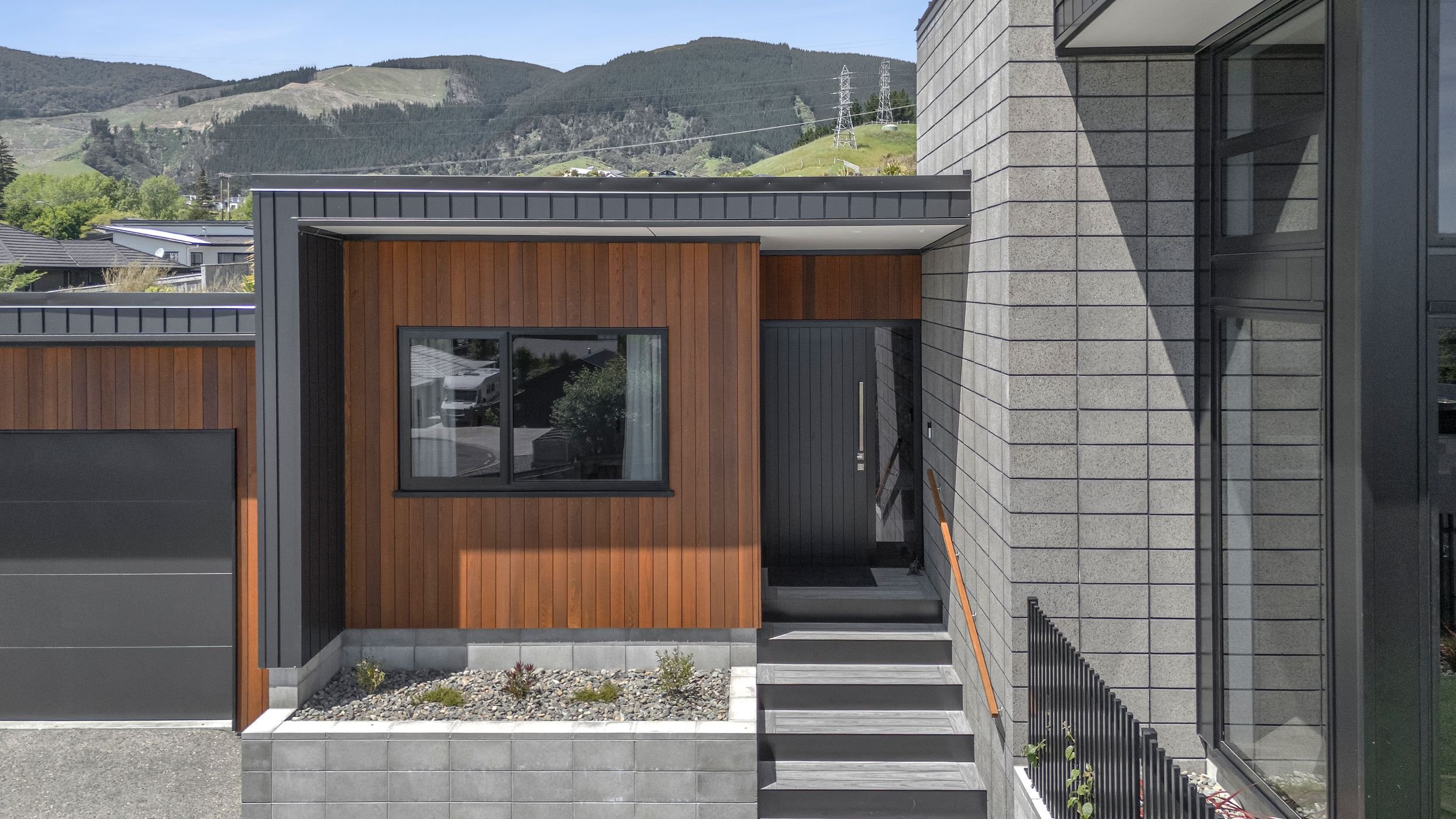 Modern house exterior with gray brick, wooden accents, large glass window, and concrete steps, with a mountainous landscape in the background.