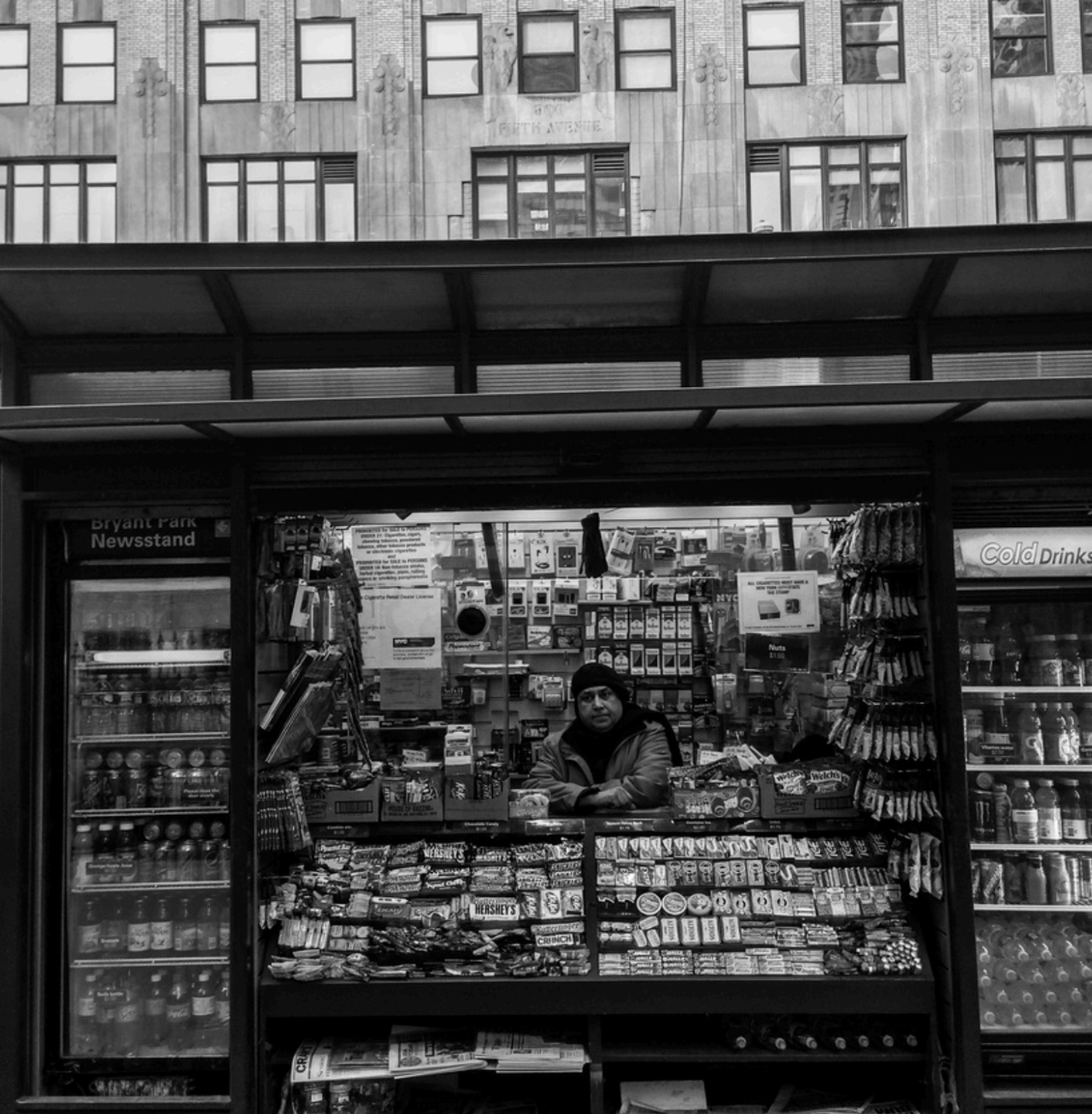 Black and White image of person sitting behind counter at outdoor vendor's stand 